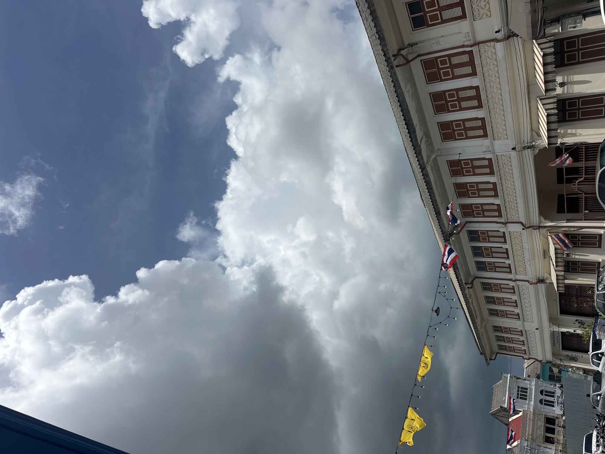 This is a medium shot, outdoor, eye-level photograph taken on a street in Phuket City Municipality, Thailand. The scene depicts a row of traditional Sino-Portuguese shophouses with white facades and dark red window shutters. The shophouses have multiple stories, with ornate decorative elements on the upper facades and awnings over some of the ground-floor entrances. Flags of Thailand are visible on some buildings. String lights with bulbs are hung across the street, interspersed with yellow triangular flags bearing an unknown emblem. In the background, several white buildings with white roofs are visible. The sky above is filled with dramatic, dark, and light grey clouds, with patches of blue sky showing through. The street in the foreground shows parked cars and motorcycles. The overall impression is a historic streetscape under a dynamic sky.