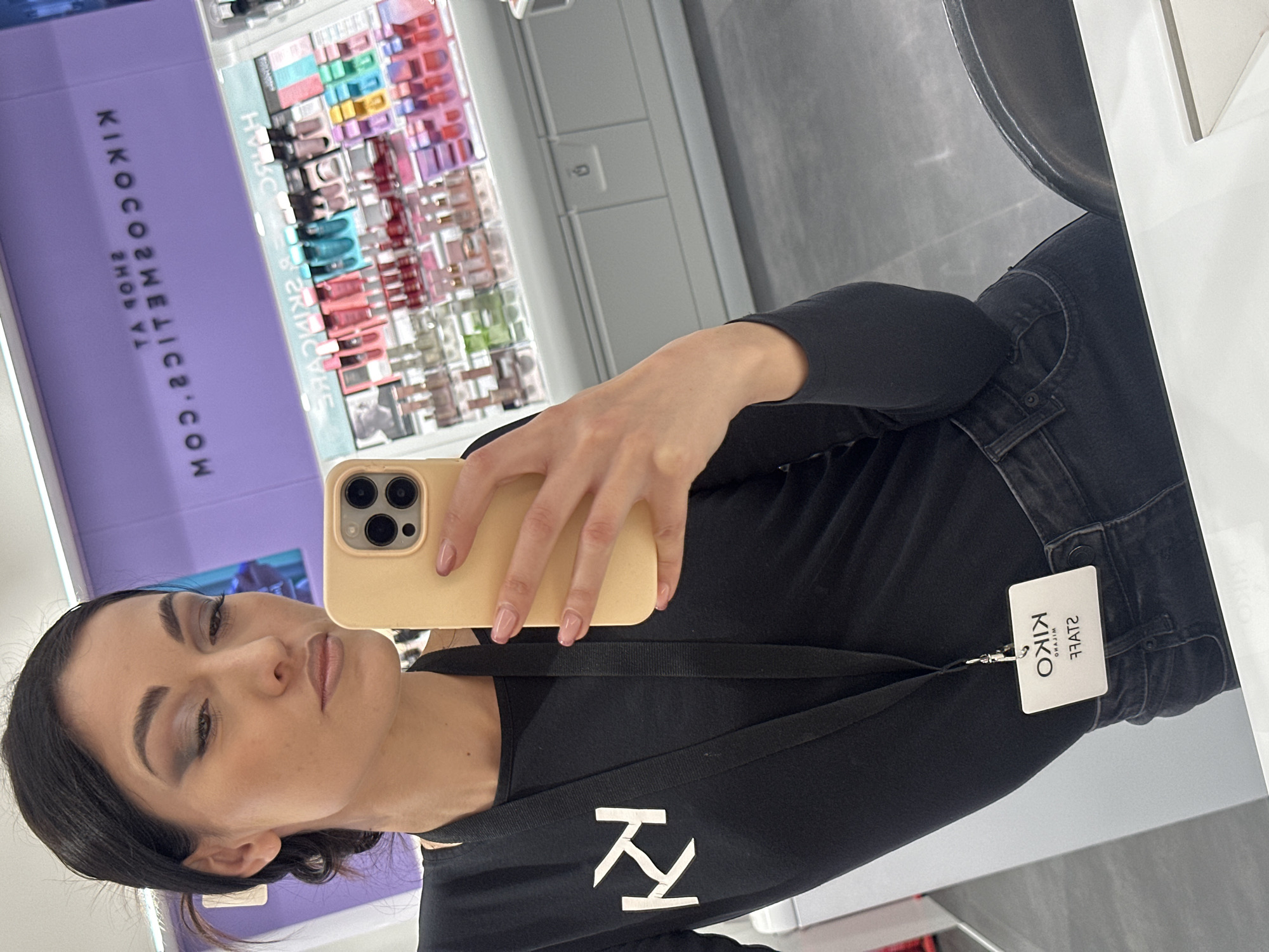 A woman with fair skin and dark hair is taking a selfie in a KIKO Milano store in an unknown city. She is wearing a black shirt with a white logo of KIKO on the front, a black lanyard, and black jeans. Her nails are painted a nude pink color. The phone she is using has a light yellow phone case. In the background, there is a display of KIKO cosmetics and skincare products. The store appears to be well-lit and clean, with light grey walls and floors. A mirror is visible to the right. The woman is wearing makeup, including eyeshadow and lipstick. She has a KIKO staff tag on her jeans. The location is in the city of Unknown, Unknown.