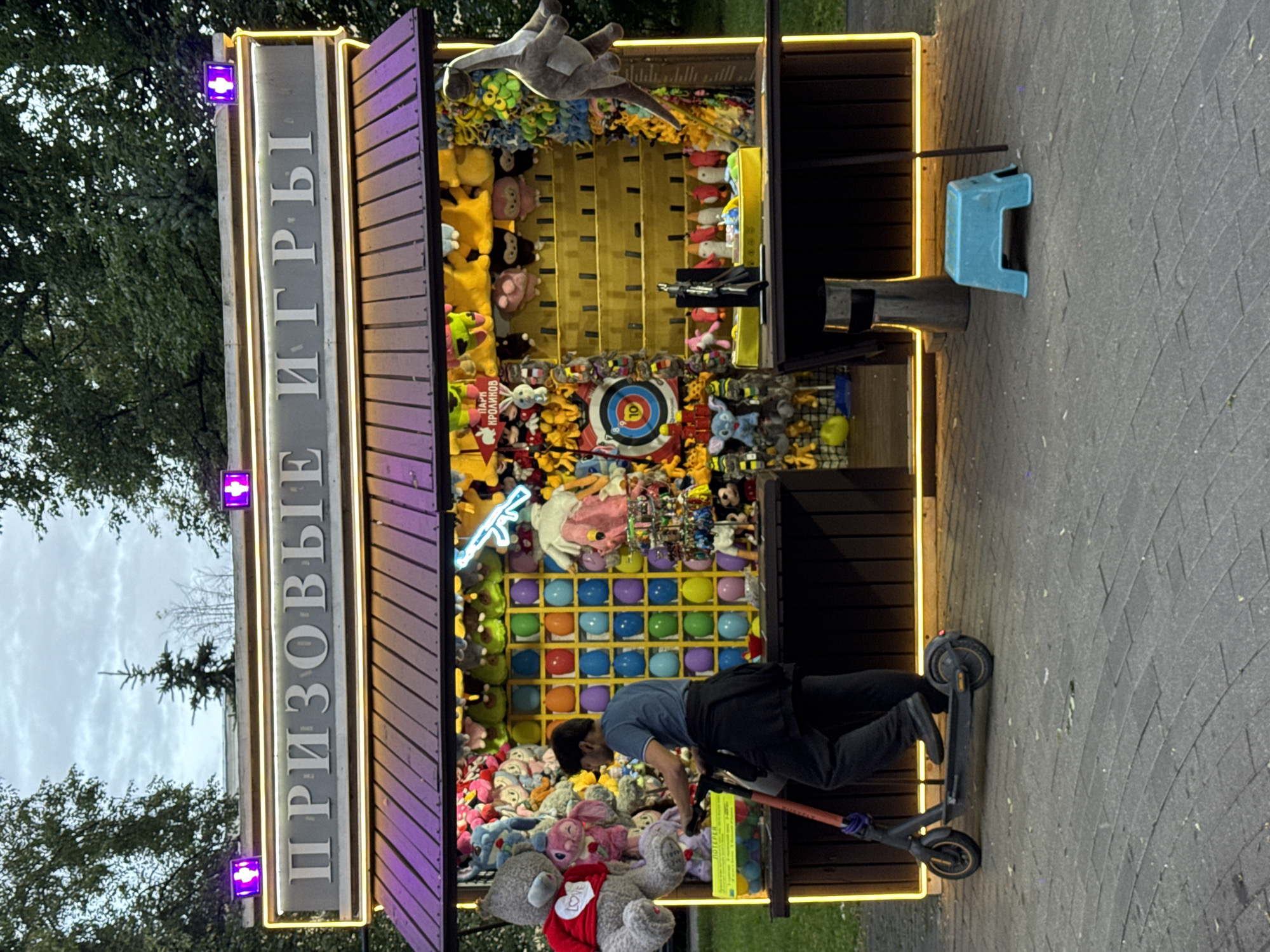 A man on an electric scooter is parked next to a carnival game stall filled with stuffed animals. The stall has a sign that reads "ПРИЗОВЫЕ ИГРЫ" (Prize Games). The man, wearing a blue shirt and dark pants, is leaning over the stall, possibly interacting with the game or prizes. The stall itself is brightly lit with neon yellow and purple lights. Inside the stall are shelves filled with various stuffed animals, balloons, and a target with darts. A blue plastic stool is placed to the right of the stall. The ground is paved with gray bricks. The scene appears to be outdoors, with trees visible in the background and an overcast sky. The time of day seems to be evening or late afternoon, given the artificial lighting.