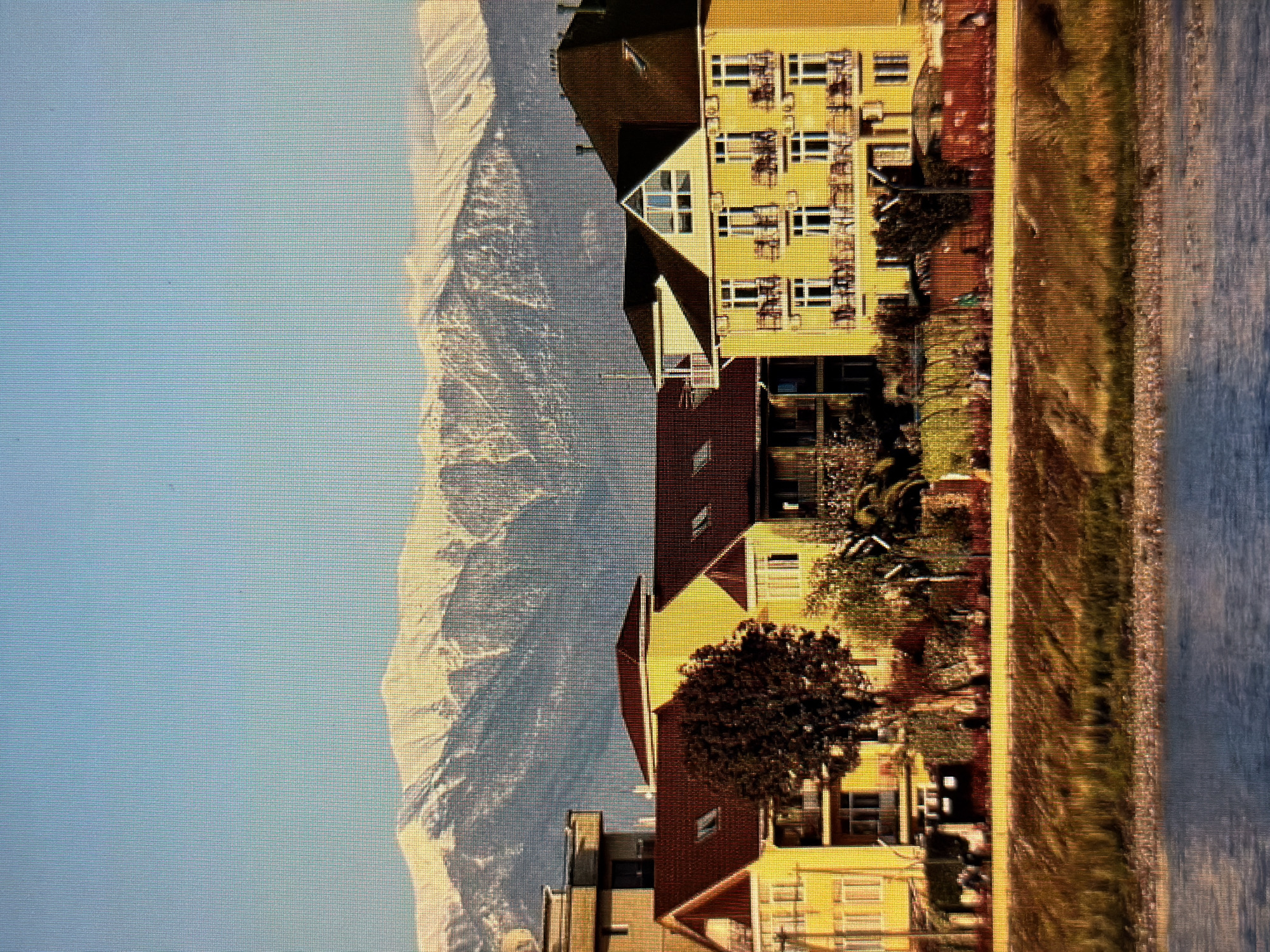 The image depicts a sunny day in Sirius, Russia, with a clear blue sky above snow-capped mountains in the distance. In the foreground, a body of water is visible, with a grassy embankment beyond it. A row of bright yellow buildings with dark red roofs lines the embankment. These buildings appear to be hotels or apartment complexes, with multiple balconies and windows. A few people can be seen walking along a path in front of the buildings, and some trees and foliage add greenery to the scene. The overall impression is one of a coastal resort town situated against a dramatic mountain backdrop.