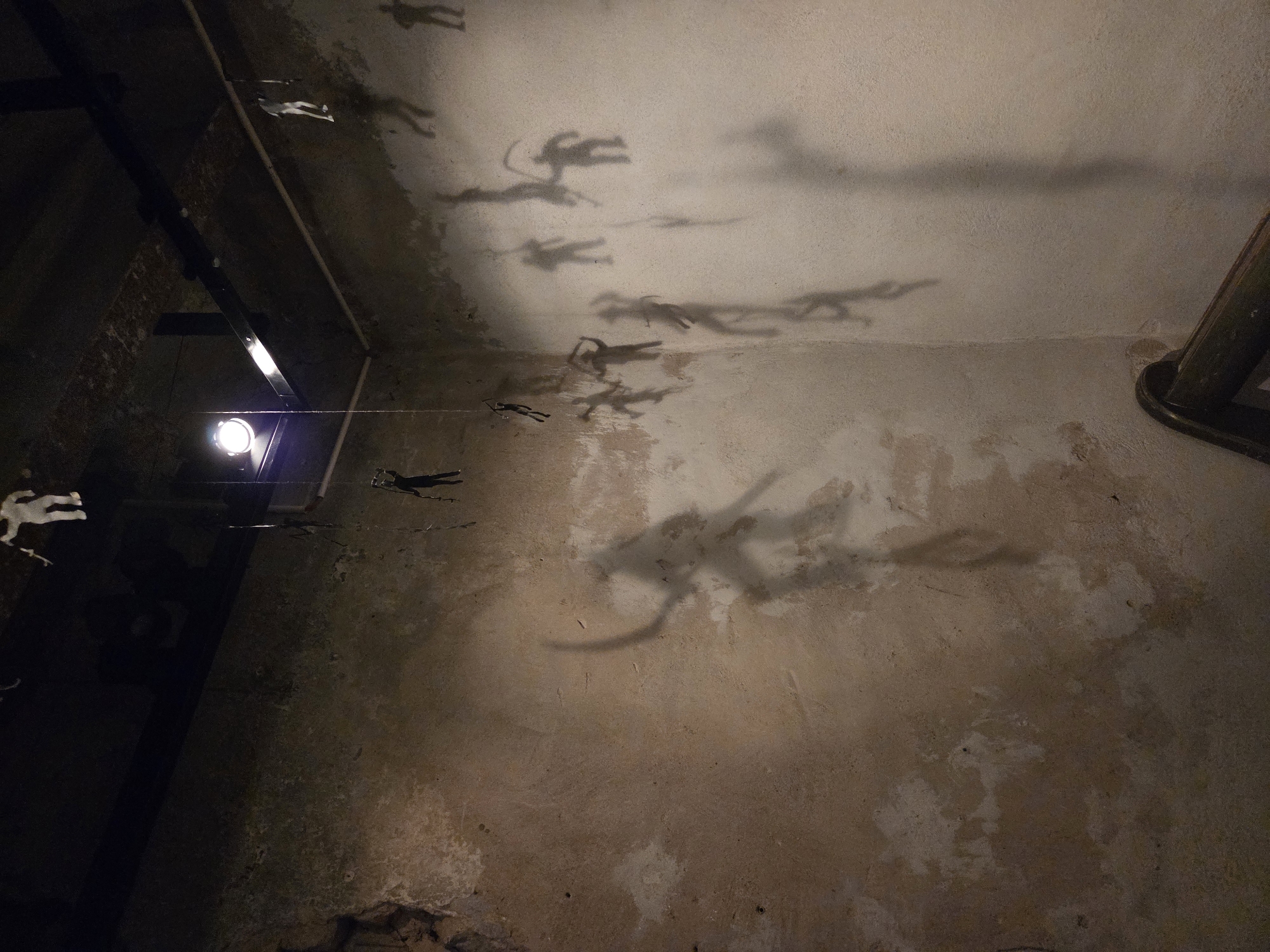 This is an interior photograph of a dimly lit corner space, possibly an art installation or a historical site. The walls are rough and textured, with patches of discoloration and wear, suggesting an aged or rustic environment. The ceiling is dark, with visible beams and track lighting.
The primary subjects are numerous small, human-shaped figures, both physical and as shadows cast on the walls. These figures appear to be suspended by thin wires from the ceiling, creating the illusion of them floating or climbing. Some figures are white and more distinct, while others are darker, appearing as shadows or silhouettes. Many of the figures are in dynamic poses, some holding what appear to be weapons or tools, suggesting a narrative of action or struggle.
The shadows cast by these figures are significantly larger and more distorted than the actual objects, adding a dramatic and possibly unsettling quality to the scene. The lighting, originating from a prominent spotlight in the upper left portion of the image and other unseen sources, plays a crucial role in creating these shadows.
The space appears to be a corner where two walls meet. In the lower right, a dark, curved object, possibly a wooden railing or part of a bench, is visible, further grounding the scene in a physical space.
Based on available information about Malmö, this scene strongly resembles the interior of the **Bunker Museum Malmö**, an exhibition space housed in a former air-raid shelter. The rough, concrete walls, the sense of being underground or in a confined space, and the artistic use of figures and shadows are characteristic of installations found in such historical and repurposed locations. The bunker was built in 1943 during World War II.