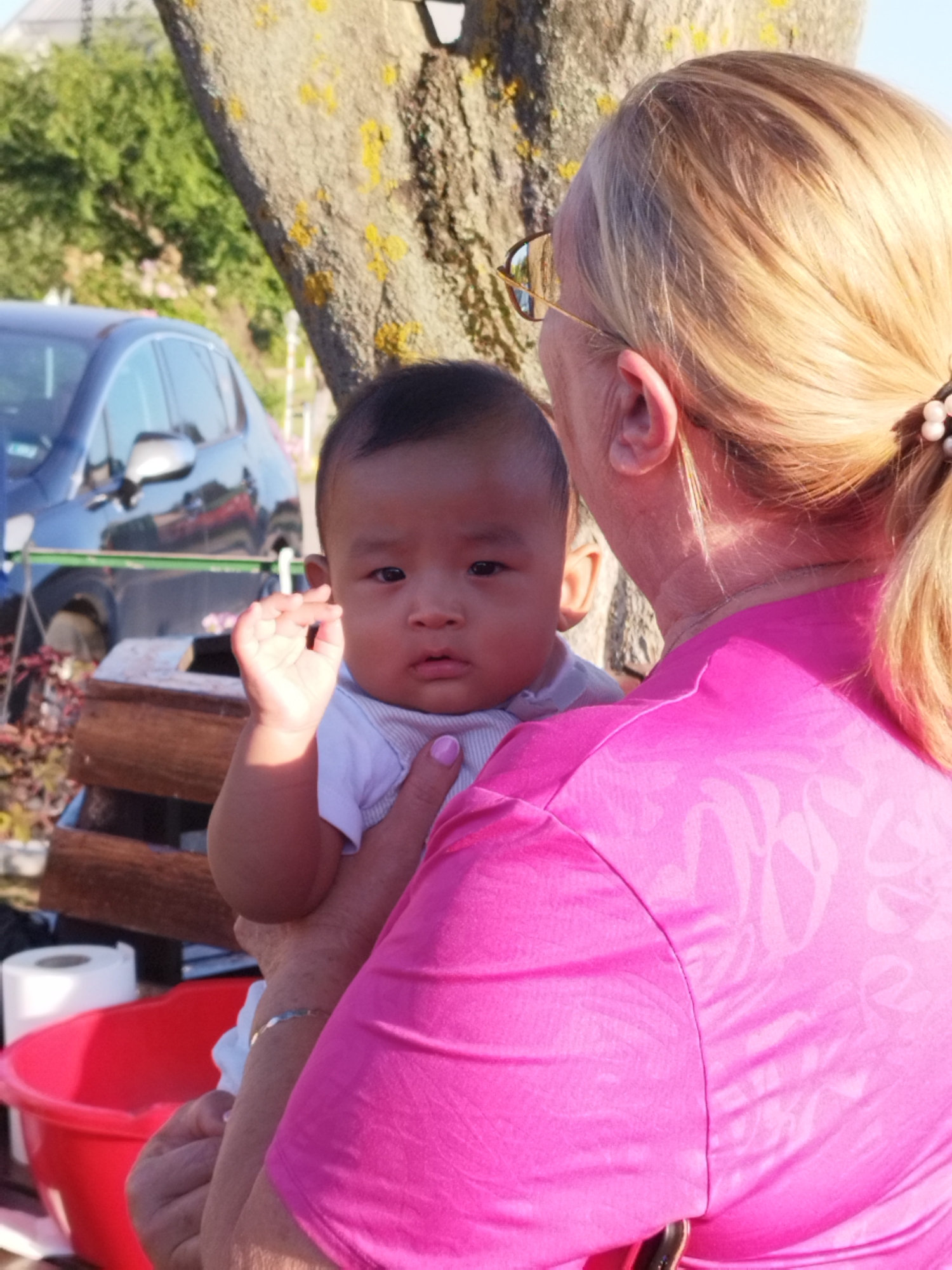 In this image, a fair-skinned woman with blonde hair, wearing glasses and a pink patterned shirt, holds a baby with dark hair and skin. The baby is wearing a white shirt and its right hand is raised. They are outdoors, with a large tree trunk and green foliage in the background. A blue car is visible to the left, and a red plastic basin is in the foreground on the right. A roll of white paper is also visible next to the basin. The lighting suggests it is daytime, possibly late afternoon. No specific details indicate the location is Bucharest, Romania.