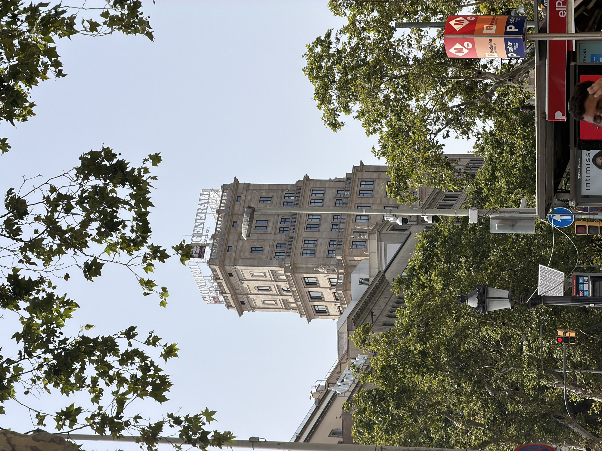 A medium shot captures the upper floors of the Edificio Generali building in Barcelona, Spain, framed by the leafy branches of sycamore trees. The building, made of stone and featuring a classical architectural style with arched windows and decorative balconies, stands tall against a clear, pale blue sky. The "GENERALI" logo is prominently displayed in large white letters on the rooftop. In the foreground, lush green trees partially obscure the view of the building's lower levels and adjacent structures. A lamppost with a solar panel attached to its base and a red traffic light are visible on the left. On the right, a signpost with the "TMB" (Transportes Metropolitanos de Barcelona) logo and "Rodalies de Catalunya" markings indicates public transport information. The overall scene suggests a sunny day in an urban environment with elements of both architecture and nature.