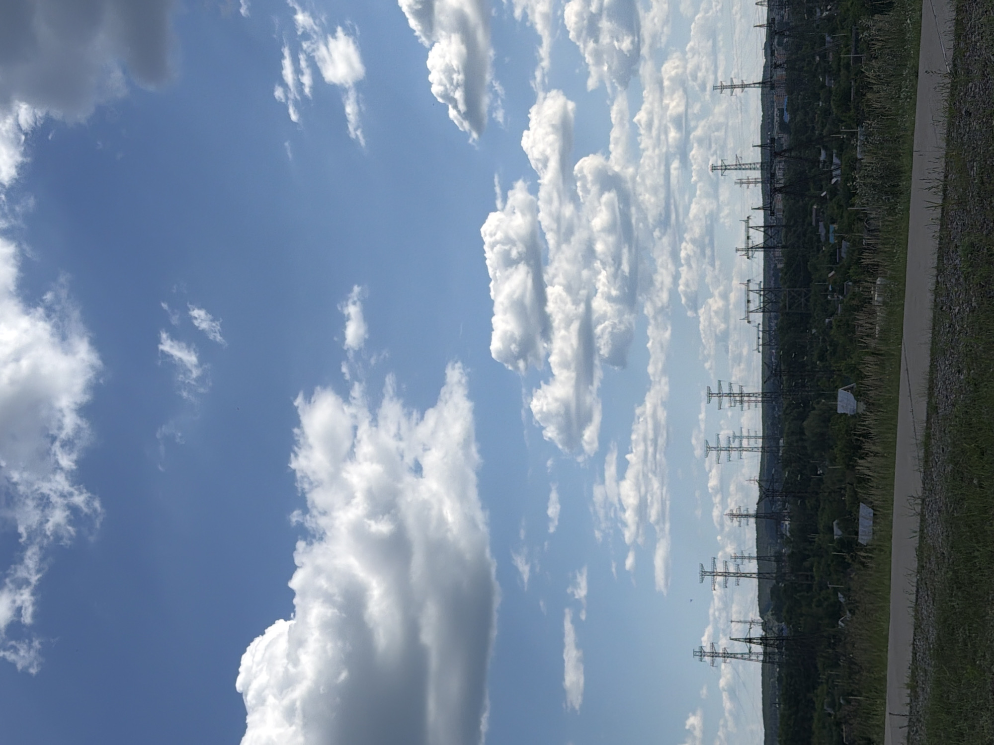 A wide shot captures a slightly elevated view of a rural or suburban landscape under a partly cloudy sky. The foreground consists of a grassy verge and a gravelly pathway. In the mid-ground, a densely wooded area is dotted with small houses, some with visible roofs, and interspersed with numerous tall, dark electrical transmission towers. The towers, some with multiple crossbars, dominate the horizon line, suggesting a significant electrical infrastructure. The background shows a distant, hazy line of more buildings and hills, indicating the presence of a settlement or city on the horizon. The sky is a vibrant blue, filled with a variety of white and gray cumulus clouds, some appearing fluffy and others more elongated. The overall scene conveys a sense of open space and a blend of natural elements with industrial structures.