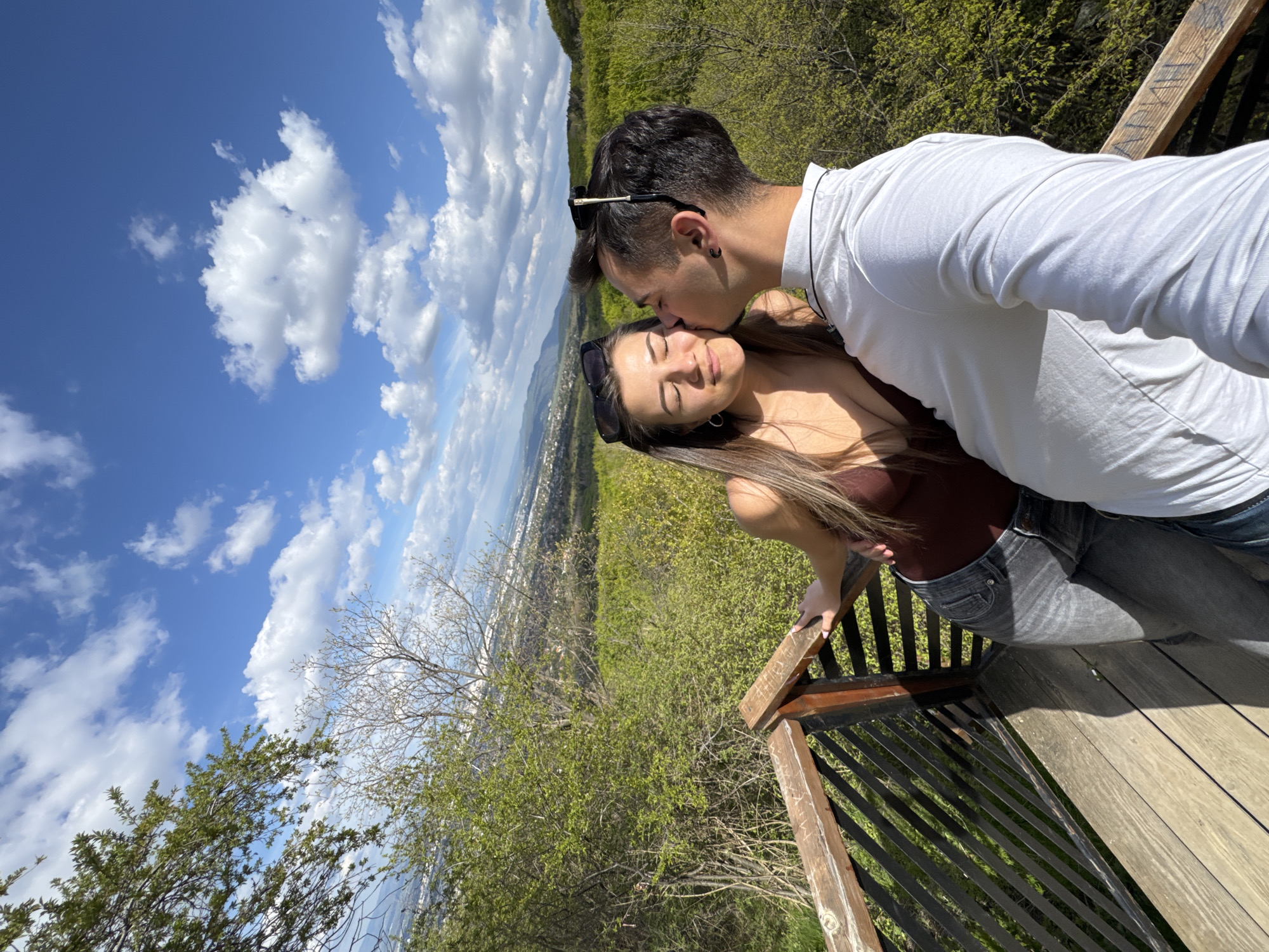 A man and a woman are on a wooden viewing platform. The man is kissing the woman on the forehead. The woman has her eyes closed. The man is wearing a white long-sleeved shirt and jeans. He has sunglasses on his head. The woman is wearing a brown tank top and gray jeans. There are trees and bushes in the background. In the distance, there is a cityscape, likely the city of Sofia, Bulgaria. The sky is blue with white clouds.