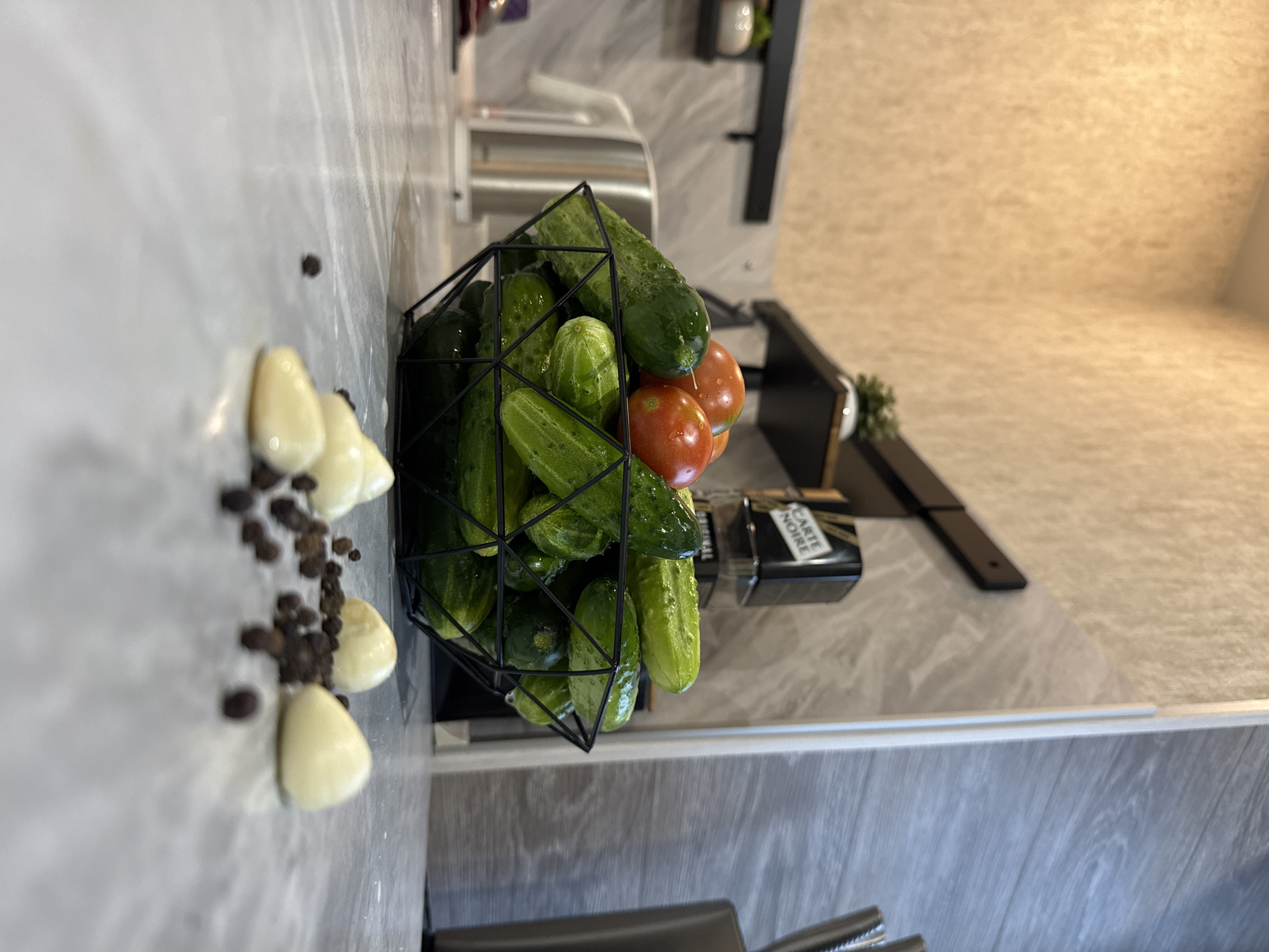 The image shows a close-up, low-angle view of a black wire basket filled with cucumbers and tomatoes, placed on a speckled gray countertop. In the foreground, several peeled garlic cloves and black peppercorns are scattered across the counter. In the background, a kettle is visible, as well as shelves with small decorative items. The overall scene appears to be a kitchen setting. The image does not contain any information that would indicate its location in the city of Солонешное, Russia.