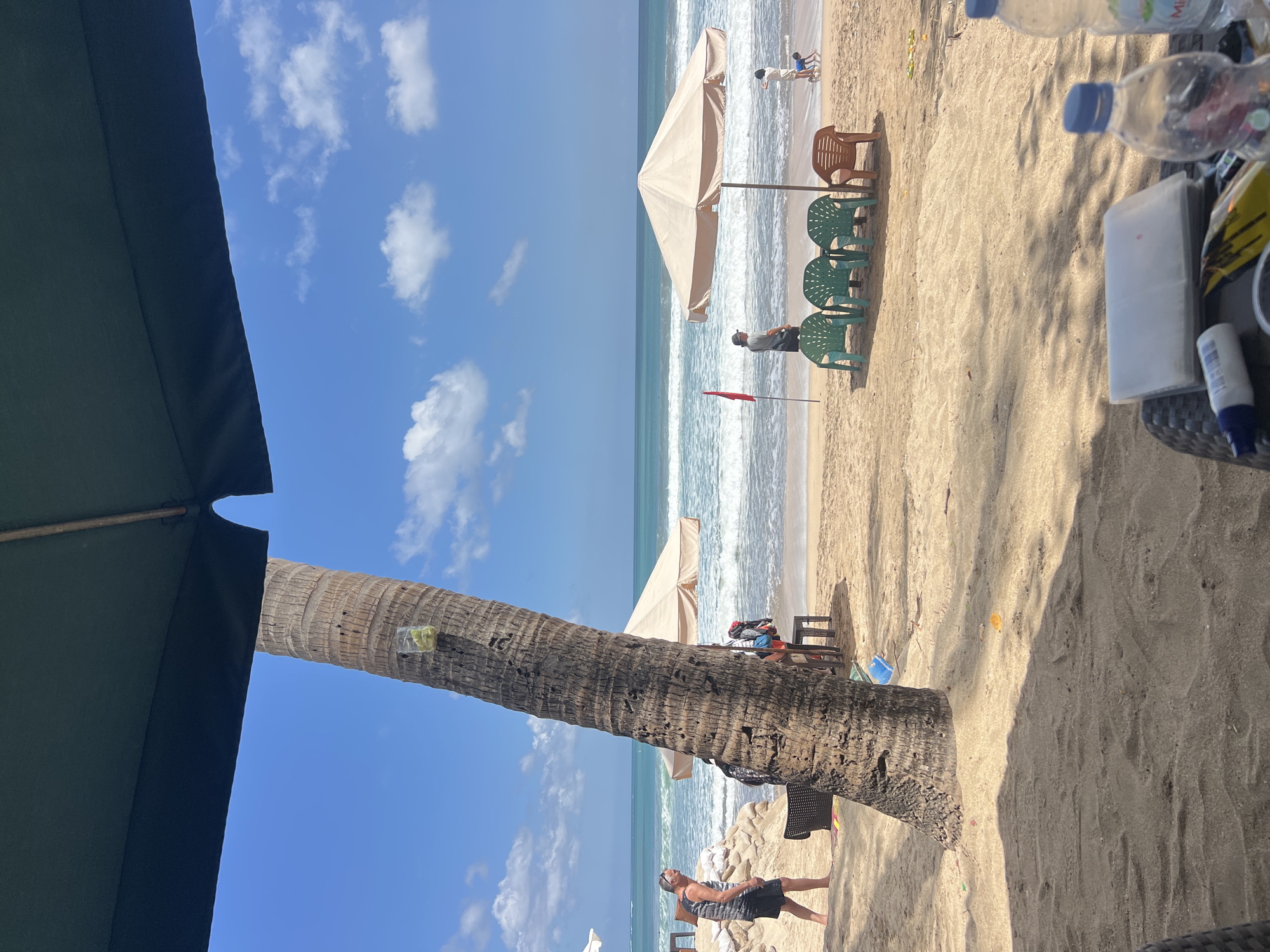 The image depicts a beach scene in Kuta, Indonesia. In the foreground, a dark green umbrella shades a sandy area with a woven table holding two plastic water bottles, one with a blue cap and a label partially visible that reads "Miracle," and a white tube, possibly sunscreen. A thin, tall palm tree trunk with a small clear cup containing green objects, likely lime wedges, attached near its top, dominates the left side of the frame.
Beyond the immediate foreground, the sandy beach stretches towards the ocean. A few plastic chairs, including three green ones and one brown one, are arranged in front of a beige beach umbrella. To the left of the chairs, a man in a striped tank top and black shorts is walking away from the viewer. Further back, near the water's edge, another man in a grey shirt and dark shorts stands facing the ocean. A red flag is planted in the sand to the left of the umbrella. In the distance, gentle waves are breaking on the shore under a bright blue sky dotted with white clouds. A small child in a blue swimsuit is visible near the water's edge to the right of the umbrella. The overall scene suggests a relaxed atmosphere on a sunny day at a beach resort.