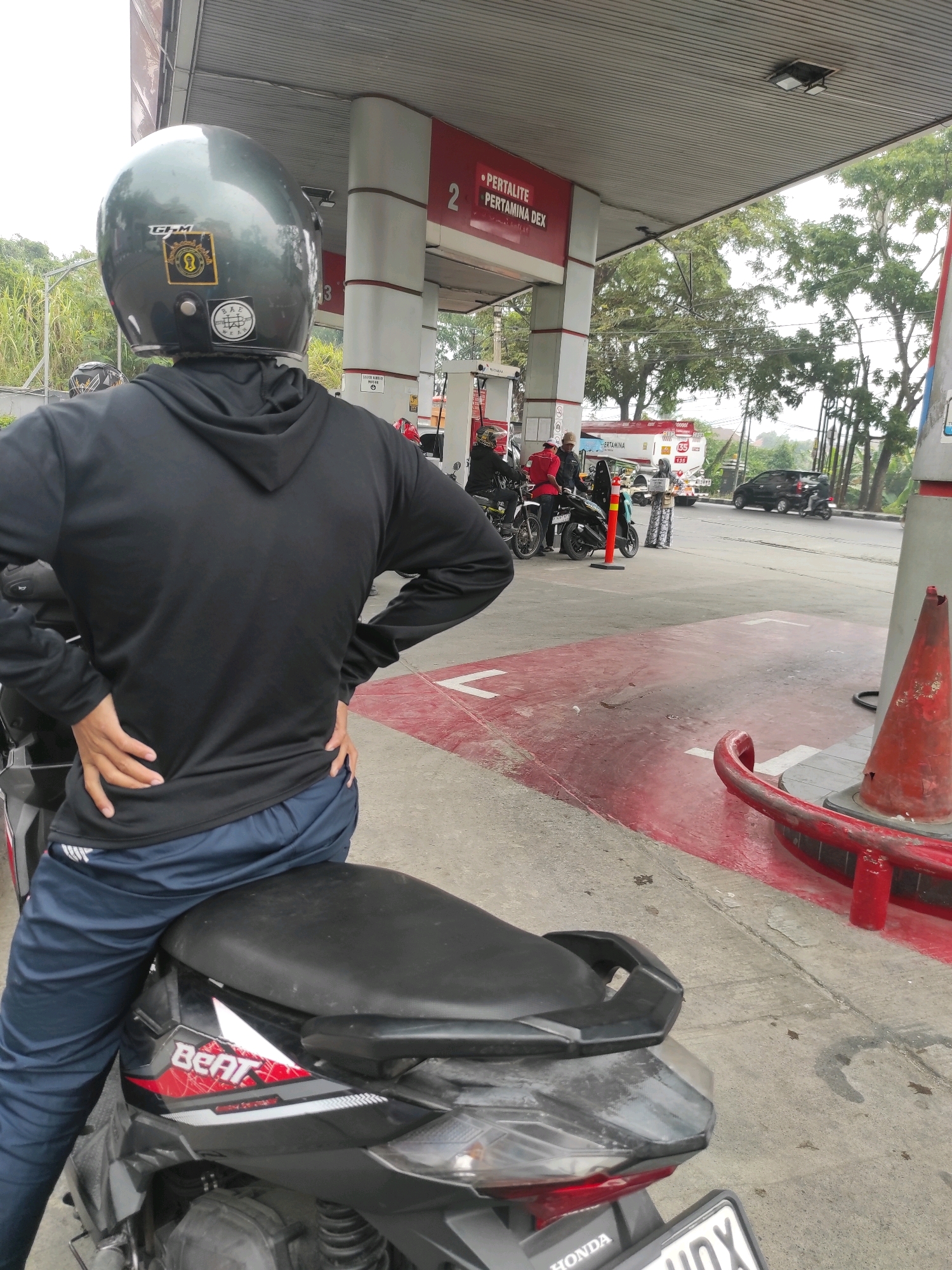 The image depicts a scene at a Pertamina gas station in Padalarang, Indonesia. In the foreground, a person is seated on a black motorcycle, facing away from the camera. The individual is wearing a grey helmet with stickers, a black hooded jacket, and dark blue pants. Their hands are placed on their hips. The motorcycle has "Beat" branding on its side.
In the background, several other motorcycles and individuals are visible near the gas pumps. One person is wearing a red uniform. A Pertamina truck with "PERTAMINA" written on its side is also present. The gas station features white and red pillars and signage indicating fuel types such as "PERTALITE" and "PERTAMINA DEX". The ground is paved with concrete and marked with red and white lines. Trees and foliage are visible beyond the gas station, along with a street where cars and motorcycles are driving. The sky is overcast. The overall atmosphere suggests a typical day at a busy gas station.