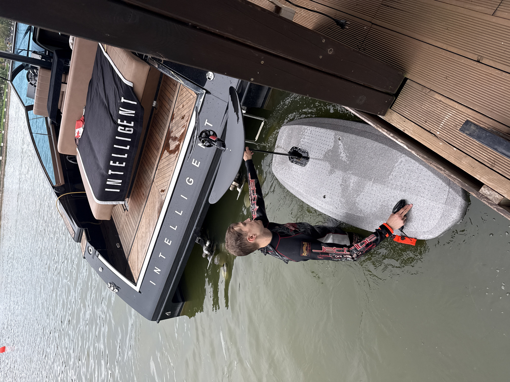 A young man in a black and red wetsuit is in the water next to a black boat. He is holding onto a wakeboard. The boat has the word "INTELLIGENT" written on a black towel draped over its seats. The boat is docked next to a wooden pier. The water is a murky green and appears to be a lake or river. The sky is overcast, suggesting a cloudy day. The scene takes place outdoors, likely in Votkinsk, Russia. The man's expression is focused as he prepares to ride the wakeboard. The overall atmosphere is one of recreational activity.