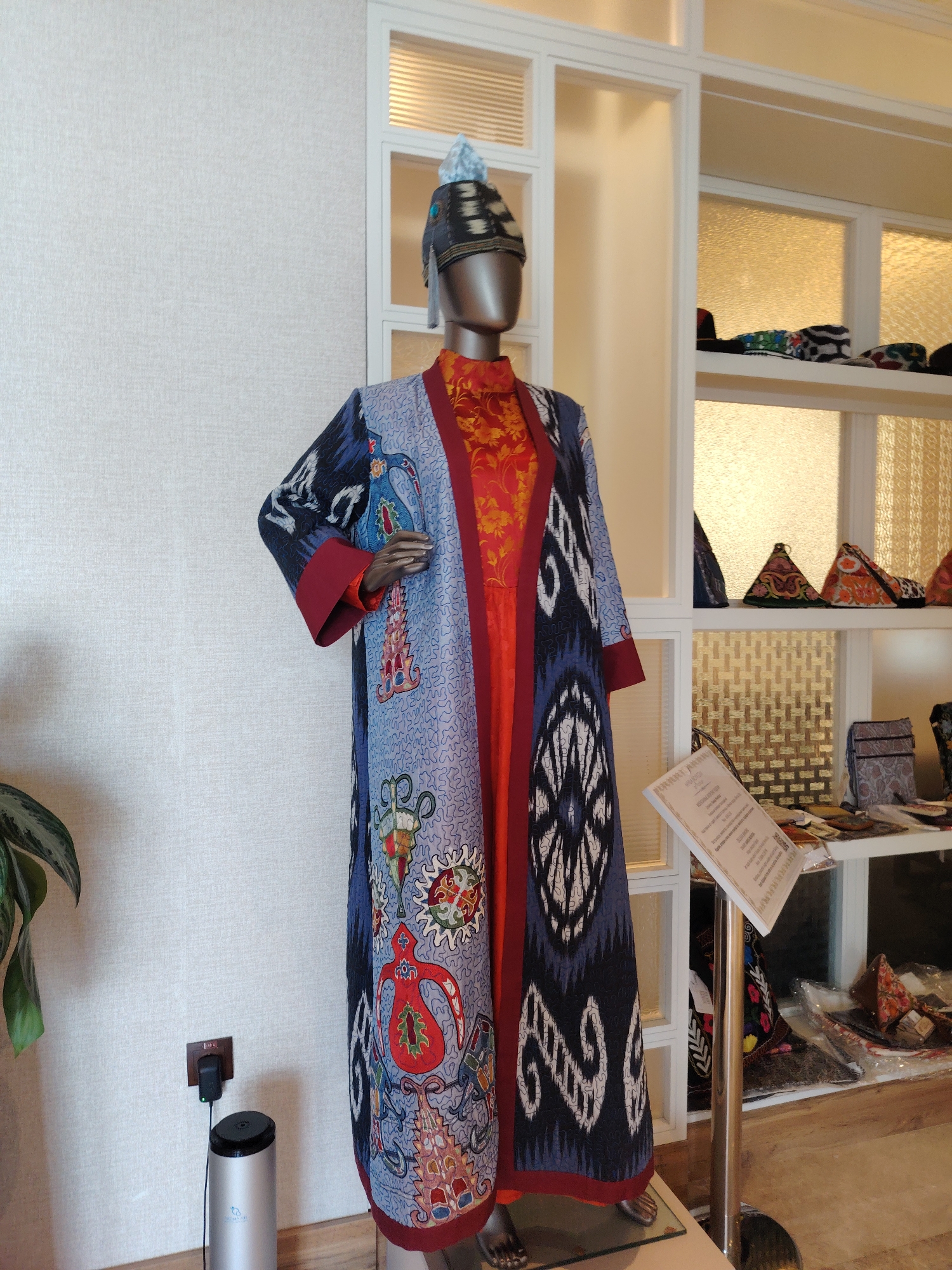 A mannequin in a traditional Uzbek robe is displayed in a brightly lit room. The robe is long and flowing, with a vibrant pattern of blue, red, and white motifs. The mannequin wears a matching patterned hat. In the background, shelves are visible, displaying various textiles and bags, suggesting a shop or museum setting. A sign stands to the right of the mannequin, providing information about the display. The room has white walls and a white framed window with translucent glass panels that emit a warm, yellow light. To the left of the mannequin, a green plant is partially visible. A power outlet and a charging device are seen on the wall behind the mannequin. The floor appears to be made of wood. The overall impression is one of cultural display and artisanal craft.