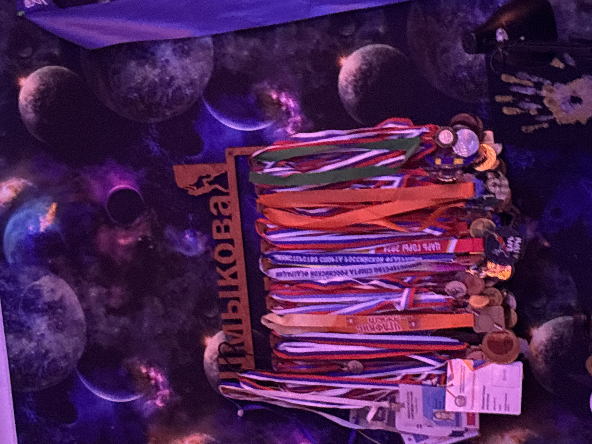A collection of medals and documents is stacked on a wooden object labeled "ПРИКАМСКИХ" (PRIKAMSKIKH). The medals are of various colors and designs. The documents appear to be identification or certification cards. The setting appears to be in a room with a dark, possibly space-themed wallpaper featuring planets and nebulae. The city is Прикамский, Russia.