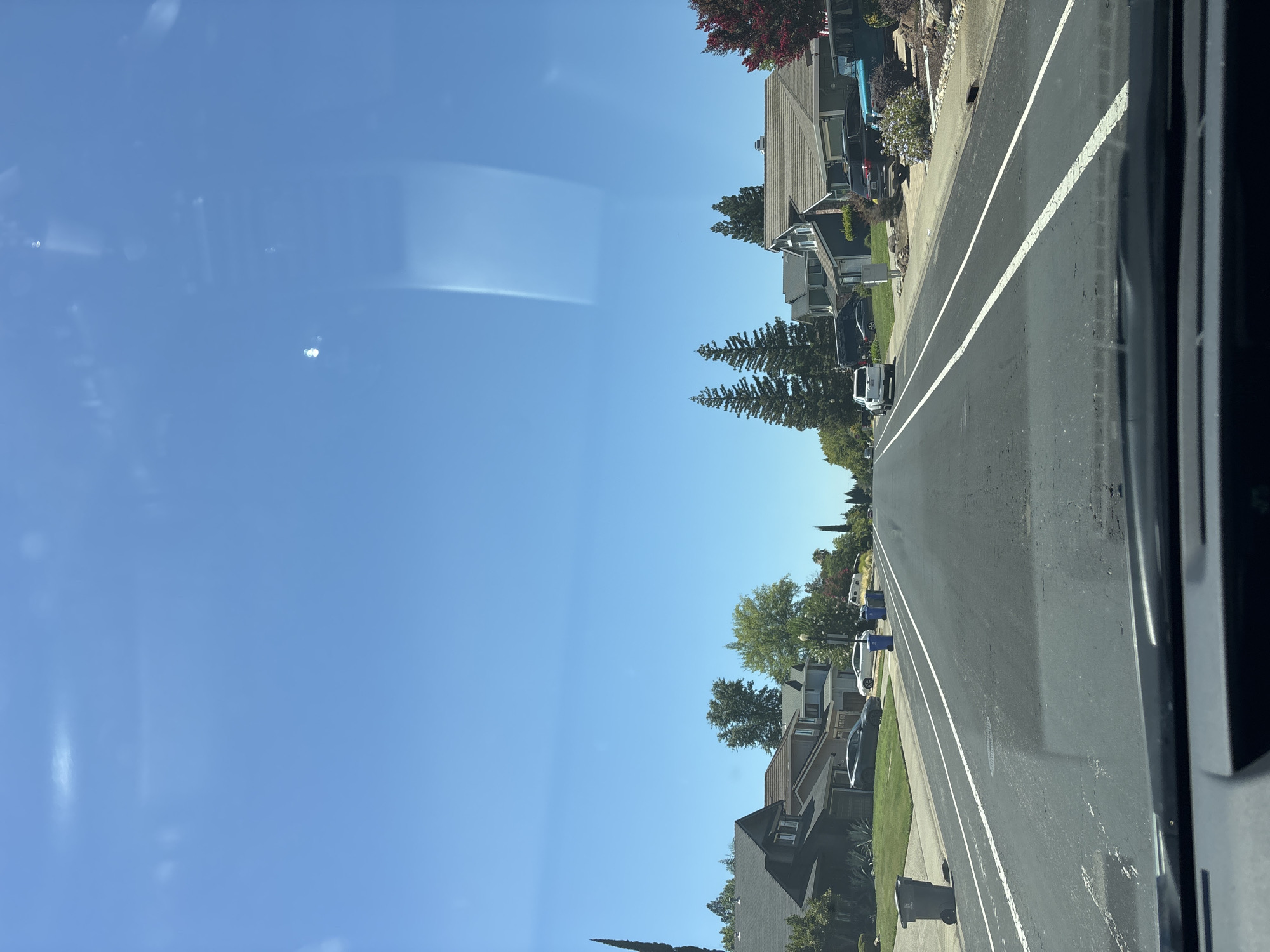 The image captures a suburban street scene in Folsom, California, under a clear blue sky. The perspective is from inside a vehicle, looking forward down a paved road lined with residential homes and manicured lawns. Several cars are parked in driveways and along the street. Trash cans are visible at the edge of the road. Trees, including tall cypress-like evergreens and deciduous trees with green foliage, are interspersed among the houses. The architecture of the homes appears to be typical of suburban American neighborhoods, with various styles and color palettes. The overall impression is one of a quiet, sunny day in a residential area.