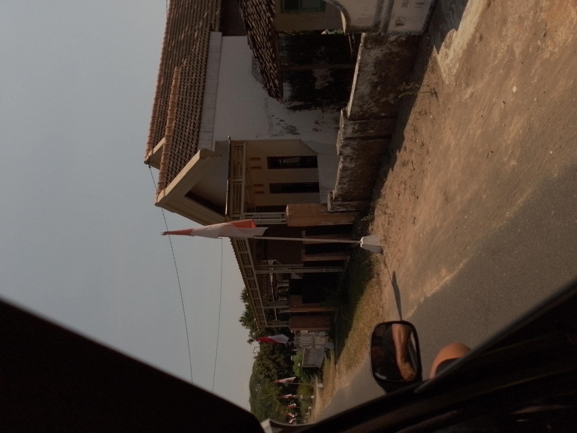 The image captures a view from inside a vehicle traveling along a paved road in Ngancar, Indonesia. The scene is dominated by a white building with a red tiled roof, characteristic of Indonesian architecture. In front of the building, a low, weathered wall lines the property, and several Indonesian flags, featuring red and white stripes, are planted along the roadside. The flags suggest a celebration or national holiday. Trees and foliage are visible in the background, indicating a semi-rural or suburban setting. The sky is clear and bright, suggesting a sunny day. The reflection in the car's side mirror shows a person's arm, implying the presence of the photographer or driver. The road itself appears dusty, with patches of dry grass and dirt. The overall impression is of a serene, ordinary day in a village in Indonesia.