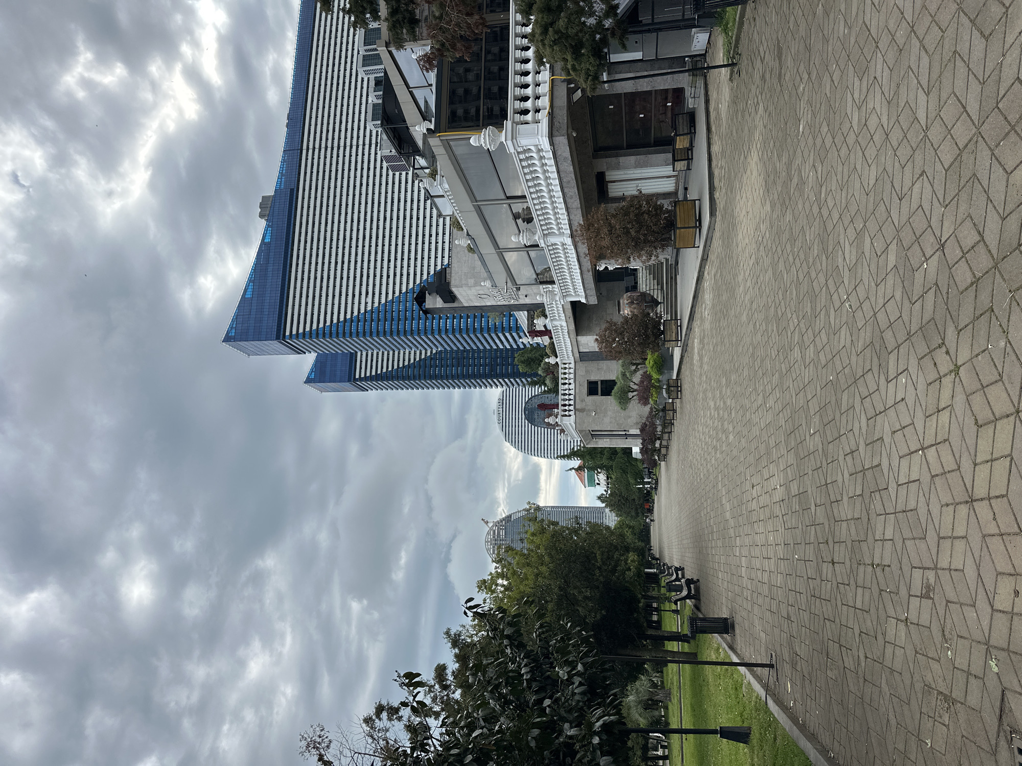 The image shows a long, paved walkway in Batumi, Georgia. On one side, there's a building with a decorative white railing. In the background, there are modern high-rise buildings with glass facades, indicative of contemporary urban architecture. On the other side of the walkway, there's a line of benches and trees, suggesting a park or promenade area. The sky is overcast with heavy clouds, creating a somber atmosphere. The patterned paving stones of the walkway are a prominent feature in the foreground.