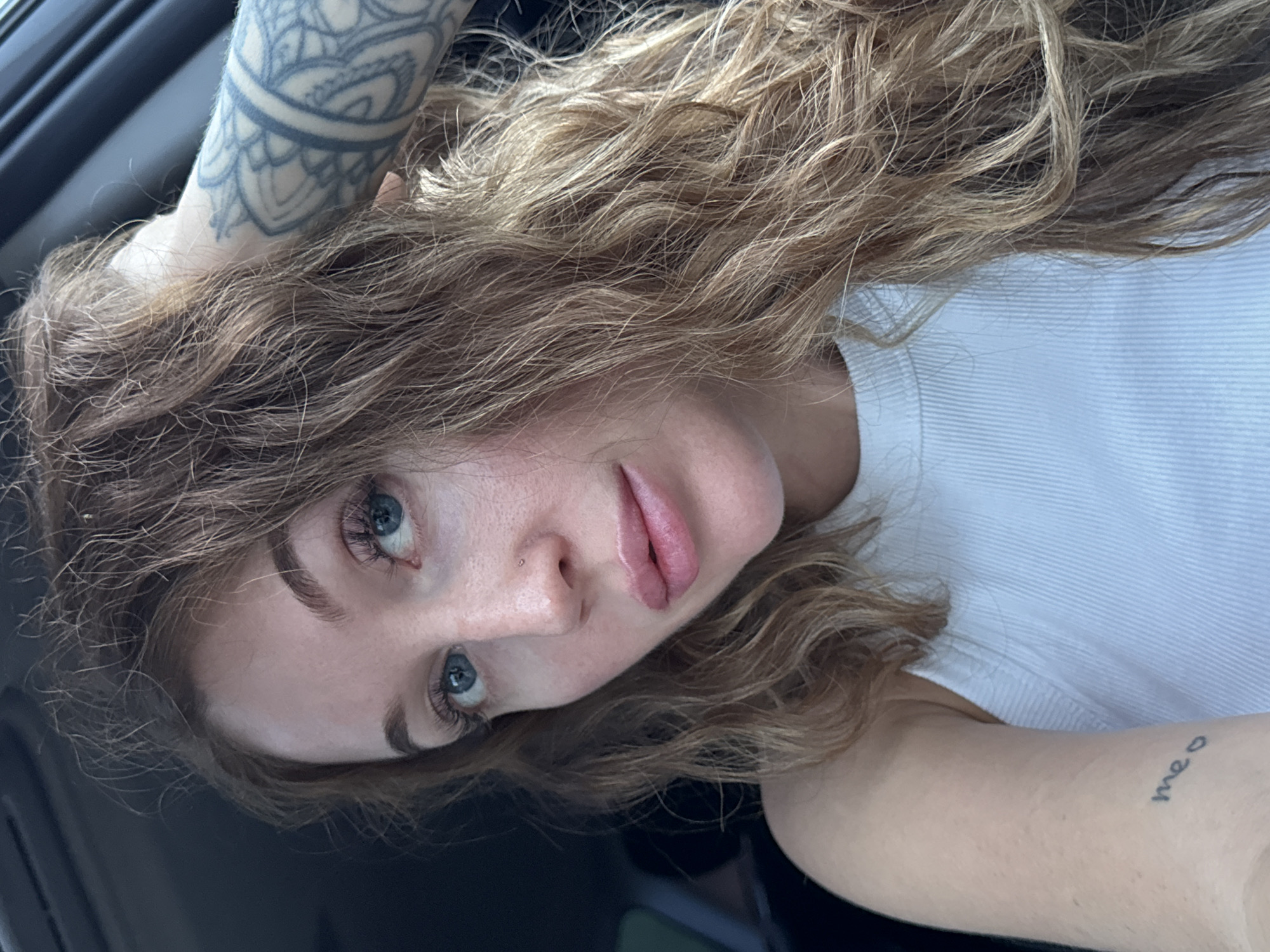 A Caucasian woman with light blonde curly hair and blue eyes is depicted in a close-up, low-angle shot. Her hair is cascading around her face and shoulders. She has a nose piercing and is wearing a white t-shirt. Tattoos are visible on both arms: an ornate circular design on her upper left arm and the word "meo" on her right forearm. The background is dark and out of focus, suggesting she is inside a vehicle. There is no evidence to suggest the location is Chaikovsky, Russia.