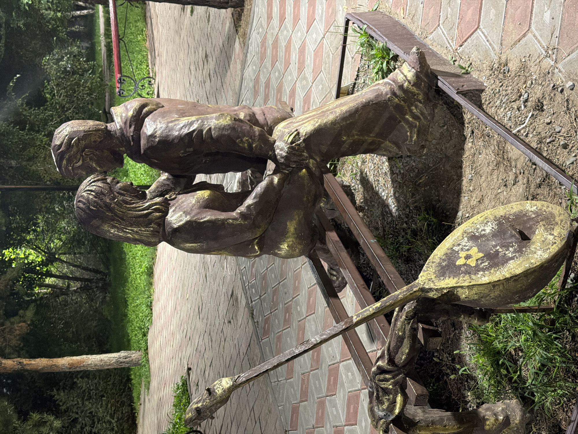 The image shows a bronze sculpture of a couple sitting together on a bench, with one figure leaning their head towards the other. A musical instrument resembling a lute or oud is placed in front of them. The sculpture is located outdoors in a park or public space with a paved walkway made of hexagonal and rectangular stones. Trees and greenery are visible in the background, along with a red bench. The lighting suggests it is evening or nighttime. This sculpture is located in the city of Pavlodar, Kazakhstan, and is part of the city's urban landscape, often found in parks or along pedestrian areas.