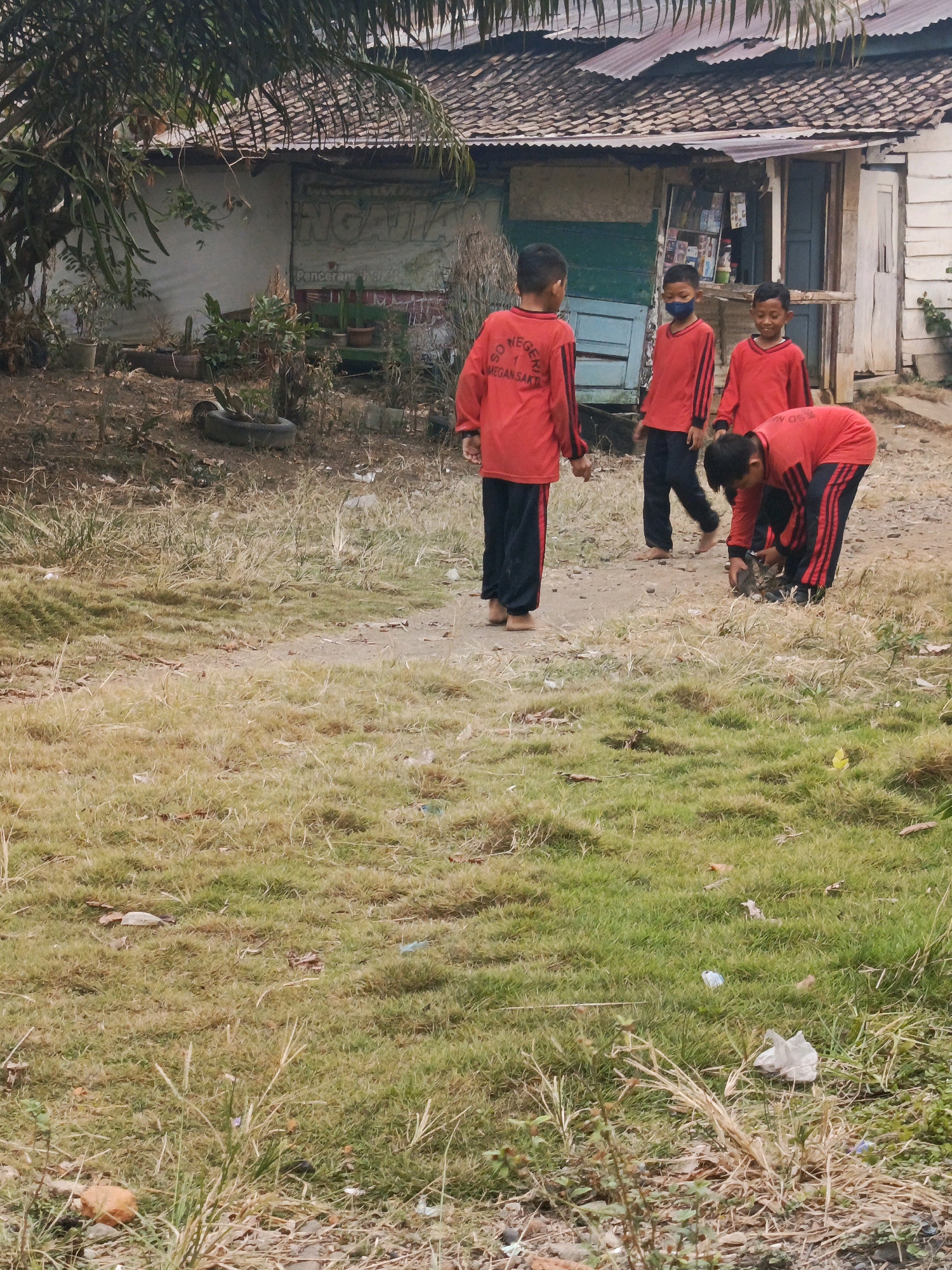 Four young boys in red uniforms with black stripes are in a yard in front of a simple building with a corrugated metal roof. One boy is standing, facing away from the camera, with the text "SD NEGERI 1 MEGAN SAKTI" visible on his back. Three other boys are also wearing similar uniforms. One boy in the center is wearing a blue face mask. Another boy is bending down, appearing to pick something up from the ground. The ground is a mixture of dry dirt and sparse, overgrown grass. The building behind them has a sign with Indonesian text and appears to be a school or small shop. The environment suggests a rural or semi-rural setting.