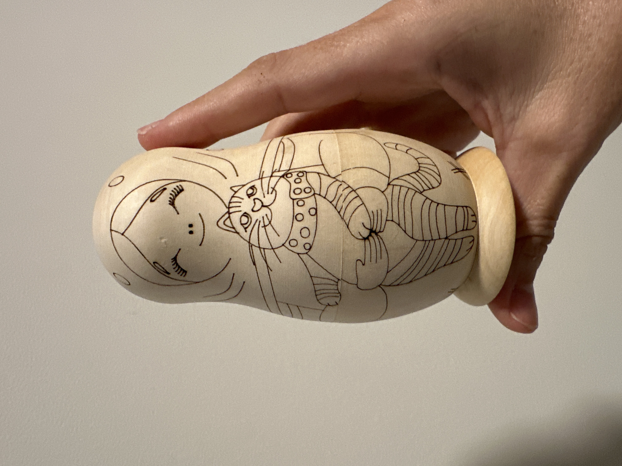 A fair-skinned hand is holding a light-colored wooden Matryoshka doll. The doll is decorated with black line drawings of a woman's face on the left and a cat with a collar and patterned body lying on top of her. The background is a plain, light gray surface. The city of Florianópolis, Brazil, is not discernible from the image.