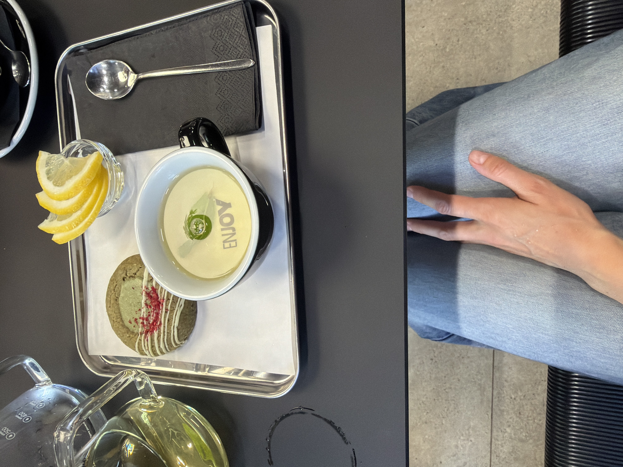 An overhead shot captures a tray on a dark surface, presenting a tea set and a cookie. On the left, a clear glass teapot with a handle and spout contains a pale yellow liquid, next to a clear glass pitcher with markings. A silver spoon rests on a black napkin to the right of the tray. In the center, a white mug with a black handle holds a light yellow liquid with a green garnish floating on top, and the word "ENJOY" is visible in white text at the bottom of the mug. Beside the mug, a cookie with white icing and red sprinkles sits on white paper. A small glass bowl filled with lemon slices is positioned above the cookie. The lower portion of the image shows a person wearing light blue jeans, with their right hand resting on their left leg. The background consists of a textured concrete floor and a ribbed black chair. The scene is set indoors, with no specific architectural features indicating a precise location within Izhevsk, Russia.