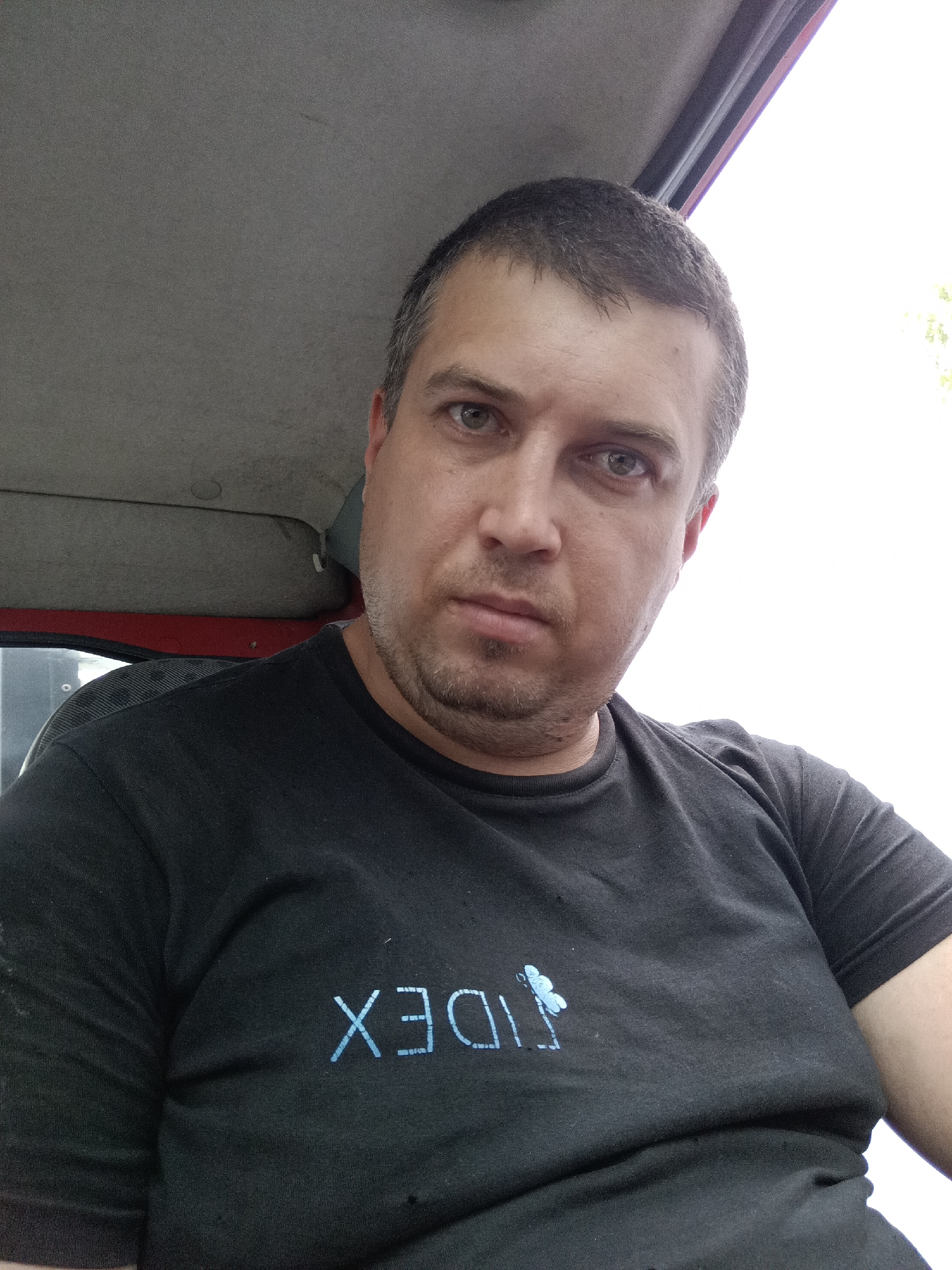 A man with short, dark hair and grey stubble is pictured in a close-up, self-taken photograph. He is wearing a black t-shirt with a light blue logo that appears to be text and a butterfly. The man is indoors, with the interior of a vehicle visible behind him, including a grey headliner and red trim. The background outside is bright white, suggesting daytime and possibly an overcast sky or a bright sun off-camera. The image was taken in Svémyslice, Czechia.