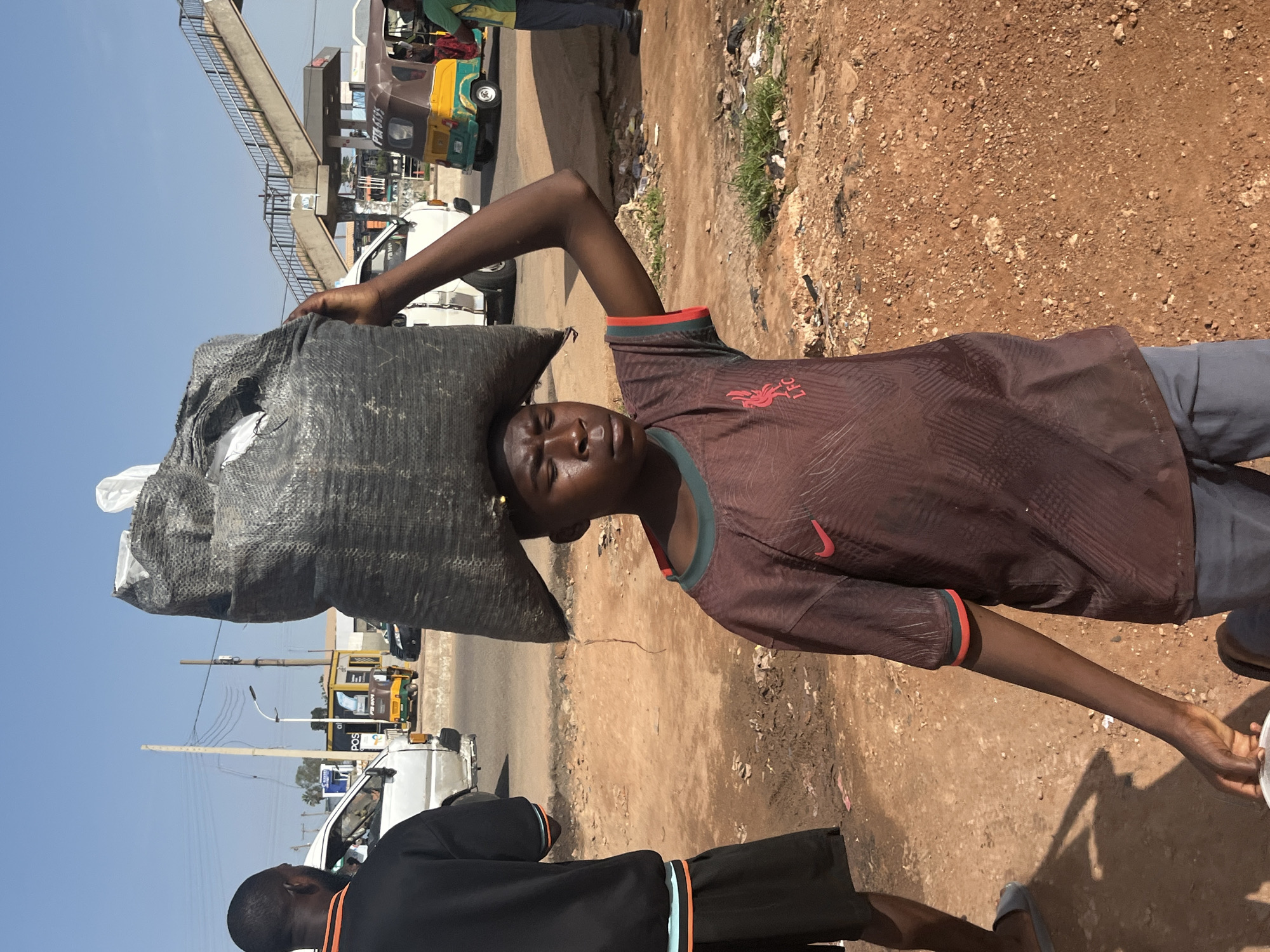 A young person is carrying a large, black, woven bag of indeterminate contents on their head. The person is wearing a brown t-shirt with a red and green trim and a red crest, and grey shorts. The person is standing on a dirt road in what appears to be a market or roadside area in Gura Top, Nigeria. In the background, there is a busy street with vehicles including a white van, a yellow and brown auto-rickshaw, and a pedestrian overpass. The sky is clear and blue. Another person, wearing a black t-shirt with orange trim and black shorts, is standing to the left of the frame, facing away from the camera. The ground is dusty and there is some litter.