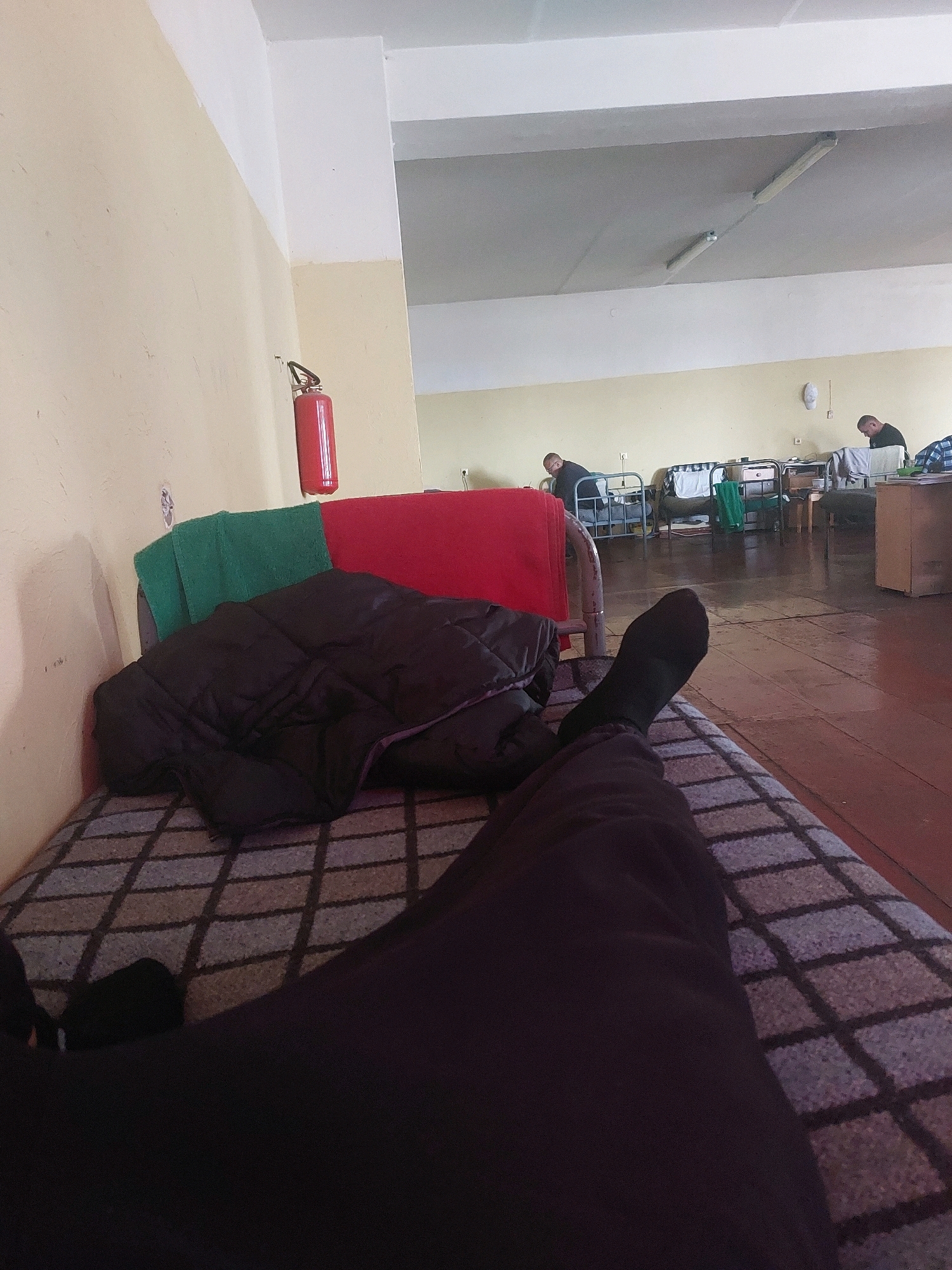 The image depicts a dormitory-style room with multiple beds lined up along a wall. In the foreground, a person is lying on a bed, with their legs and feet visible, wearing dark pants and black socks. A black quilted blanket and a red and green textile are draped over the bed. A fire extinguisher is mounted on the wall. In the background, two men are seated or leaning on beds, appearing to be engaged in individual activities. The room has a plain yellow wall and a light-colored ceiling with fluorescent lighting fixtures. The floor is made of reddish-brown planks. The overall impression is that of a communal living space, possibly a barracks or a similar institution, located in Jēkabpils, Latvia.