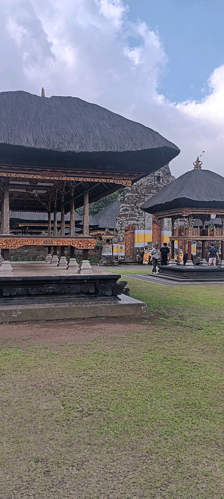 The image depicts the exterior of the Taman Ayun Temple in Baturiti, Indonesia. The temple features traditional Balinese architecture with multi-tiered roofs covered in thatch. In the foreground, a grassy lawn is visible, leading towards intricately carved wooden structures and stone bases. Several people are present, some appearing to be tourists or visitors, walking around the temple grounds. The sky is overcast with scattered clouds. The overall scene conveys a sense of cultural heritage and serene beauty.