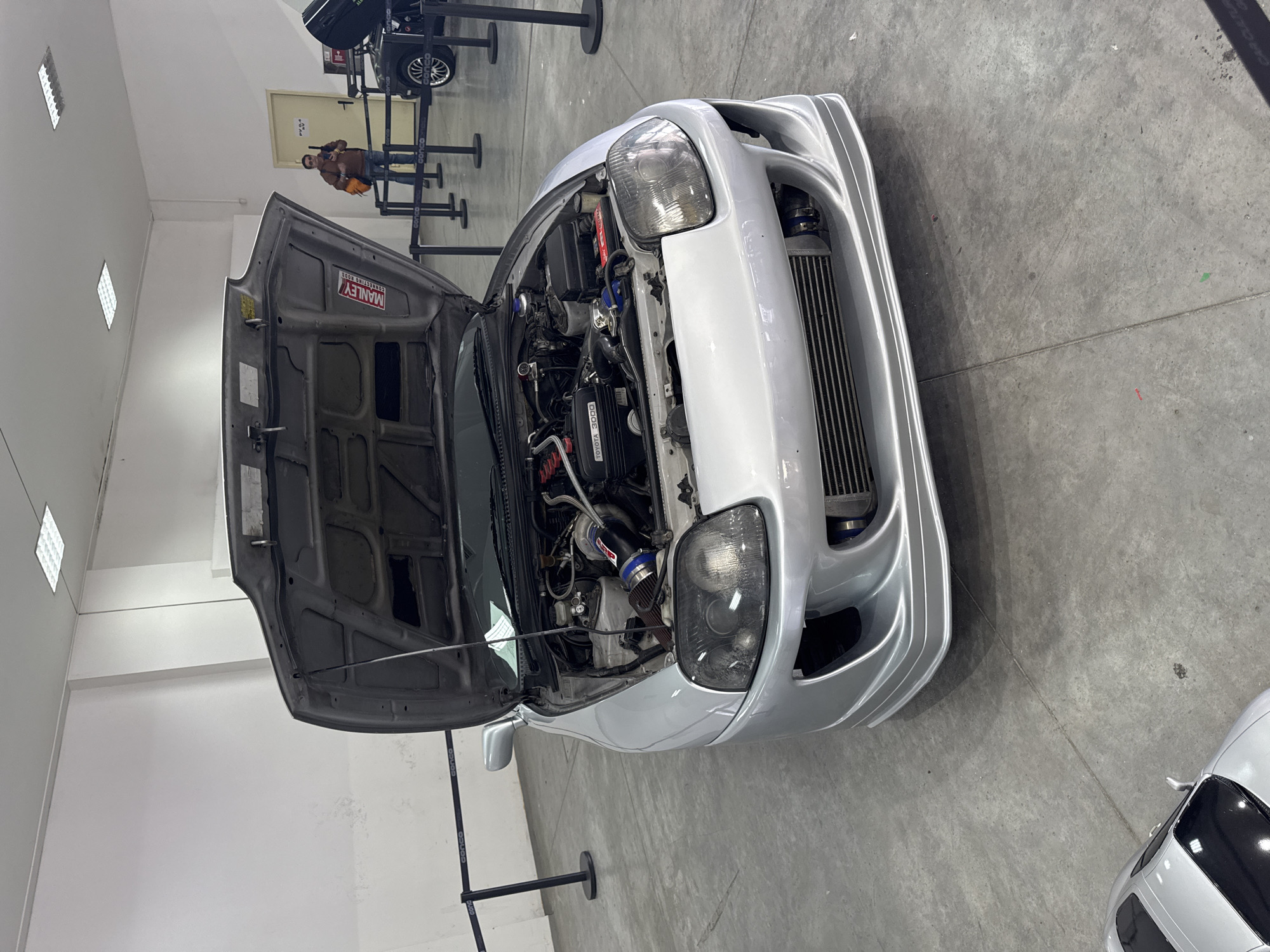 The image shows a silver Toyota Supra with its hood open, revealing the engine. The car is located indoors, likely at an exhibition or event. A person is visible in the background near a vehicle with a black cover. The image was taken in Plovdiv, Bulgaria.