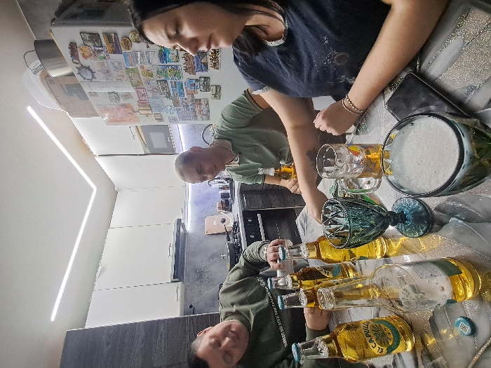 Three people are gathered around a table. Several bottles of beer labeled "El Capitan" and various glasses are on the table. One glass is full of a frothy beverage. One person is pouring liquid from a beer bottle into a glass. The setting appears to be a kitchen, with a refrigerator adorned with magnets visible in the background.