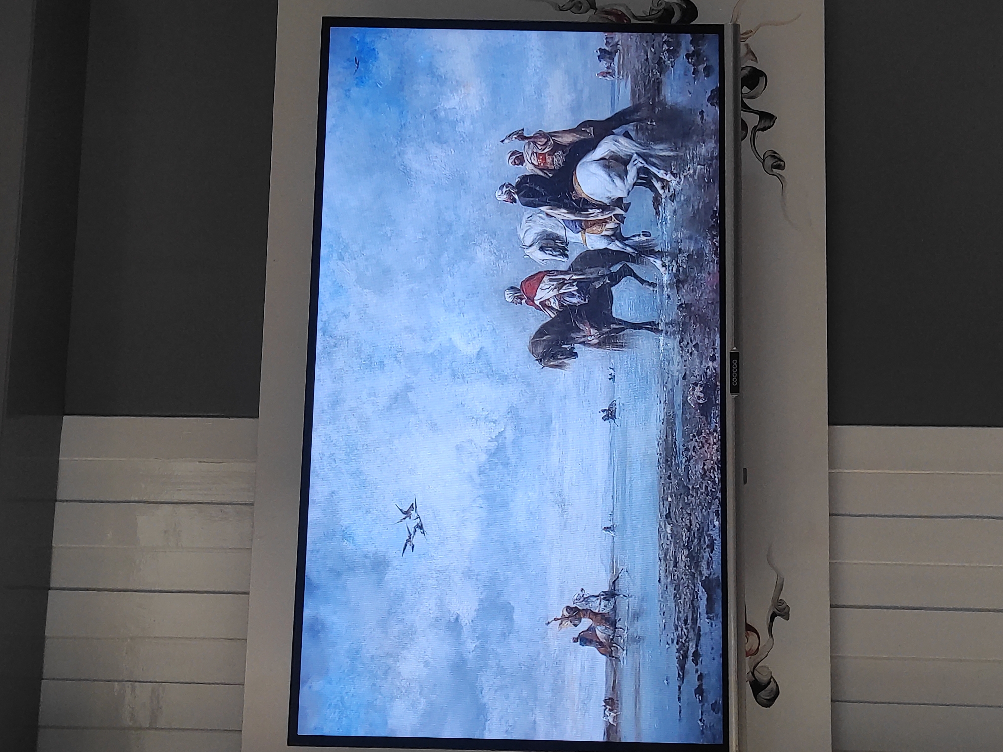 The image displays a large flat-screen television screen showing a painting of a group of people on horseback near a body of water under a cloudy sky. The painting depicts several figures dressed in what appears to be traditional Middle Eastern attire, with turbans and flowing robes. They are on horseback, with some horses white and others dark. In the foreground, the ground is rocky and wet, suggesting a shoreline or shallow water. Further back, more figures on horseback are visible, along with birds flying in the sky. The style of the painting is realistic, with detailed rendering of the figures, animals, and the atmospheric conditions.
The television is mounted on a wall which is decorated with a pattern of swirling grey and black lines. Below the television, there is a white surface with vertical lines, possibly blinds, and a grey wall beyond that. The painting itself is the dominant subject of the image, presented as if displayed on a screen.
The question asks for the location in the city of Sumedang, Indonesia. However, the image itself contains no visual cues or information that would allow for the identification of its location within Sumedang, Indonesia. The painting depicts a scene that is not geographically specific to any particular location in Indonesia, and the surrounding environment (the wall, the blinds, the TV) is generic and could be found anywhere. Therefore, it is not possible to determine the location in Sumedang based on the provided image.