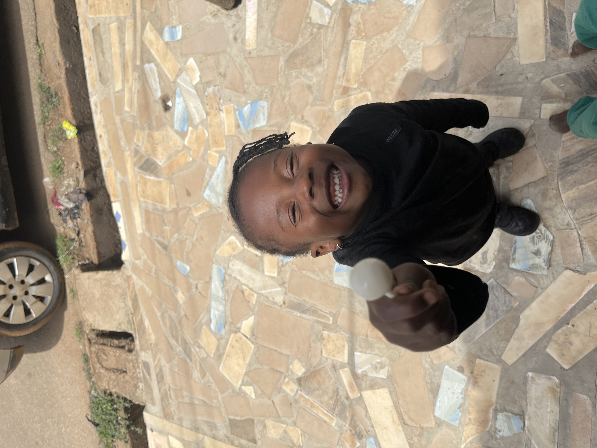 A young person with dark skin, wearing a black sweatshirt, is smiling and looking up at the camera. They are holding a white object, possibly a lollipop, towards the camera. The person has braided hair.
The setting appears to be an outdoor location with a mosaic-tiled ground. In the background, there is a vehicle with a visible wheel and a roadside area with some vegetation. Based on visual context and the prompt's specification, the location is in the city of Jos, Nigeria.