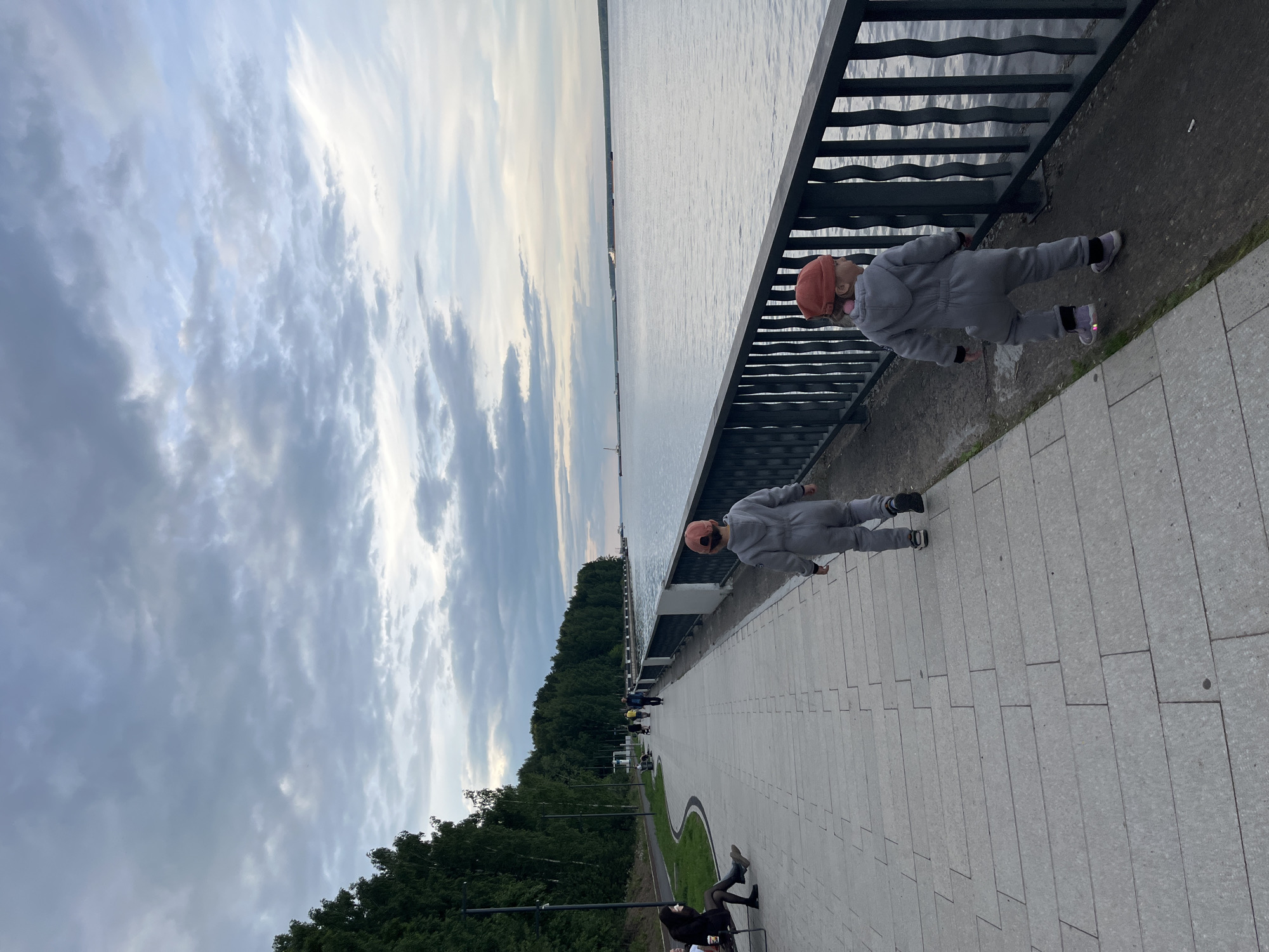 Two young children in grey jumpsuits and red caps walk along a wide, paved walkway next to a dark metal railing. The walkway borders a large body of water, likely the Kama River. The sky is overcast with dramatic clouds, and a line of trees forms the background on the left. Several other people are visible further down the walkway, and some are sitting on benches. The scene appears to be the embankment in the city of Chaikovsky, Russia, a popular area for recreation.