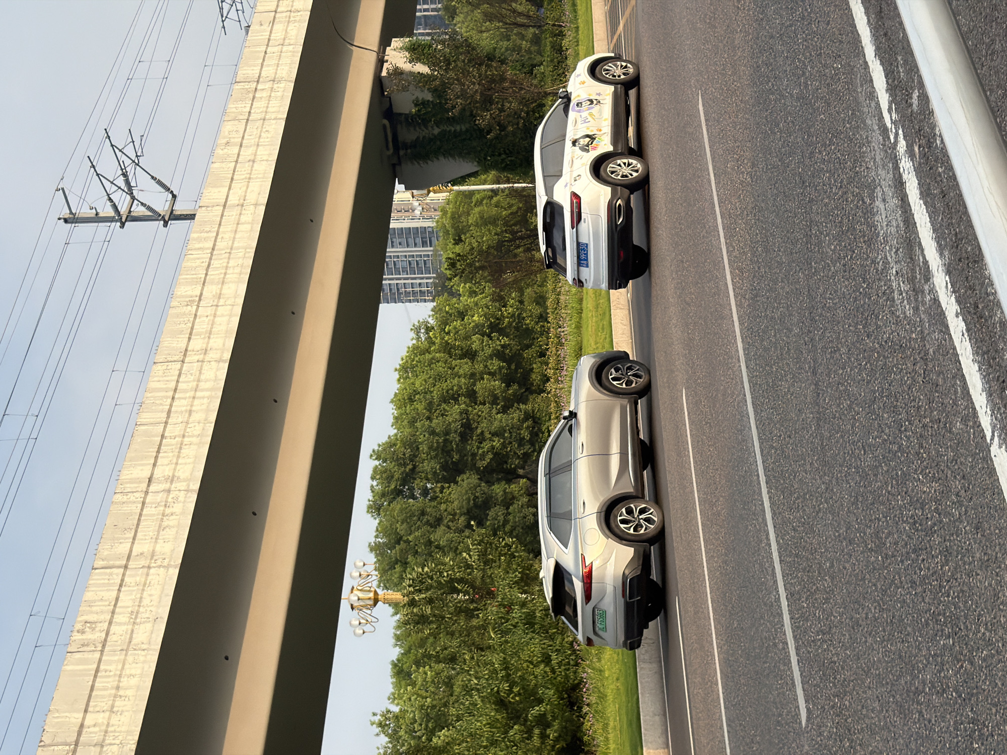 A silver SUV is parked on the right side of a road, under an elevated concrete structure, with a white SUV parked next to it. The silver SUV has license plate "E FG583". The white SUV has cartoon stickers on its side and license plate "A 9PE30". The road has white lane markings. Trees and buildings are visible in the background. The image is taken from a low angle, with the sun casting shadows from the elevated structure. The sky is clear and blue. The scene appears to be in a city. The date and time are not discernible from the image.