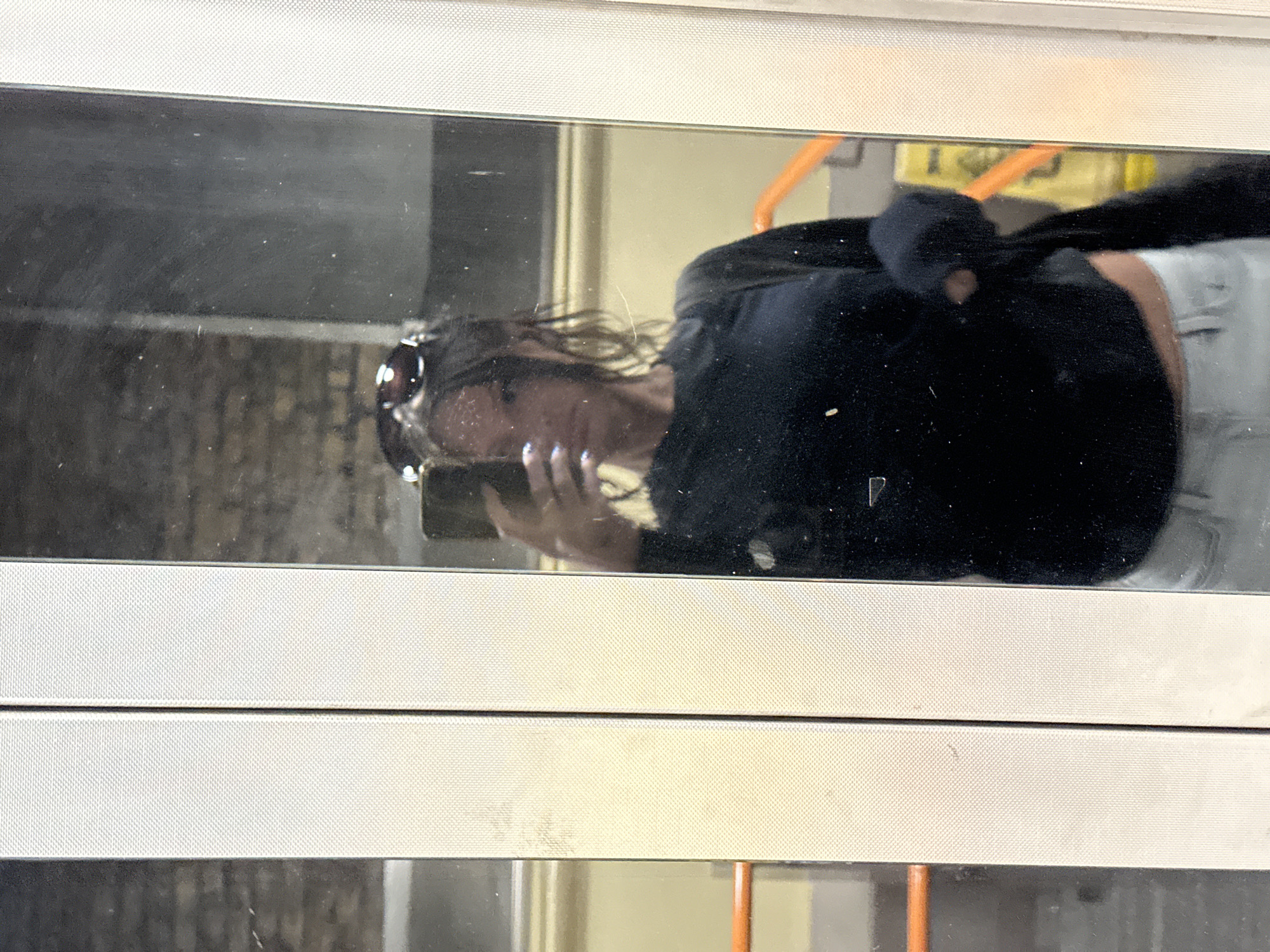 The image, originally rotated 90 degrees clockwise, depicts a reflection of a person, likely a woman, on a dark, smudged, and dusty reflective surface. The reflection is framed by two prominent light-colored, textured horizontal panels, possibly part of a window or display frame.
The central subject, seen in reflection, is a person with long, dark, wavy hair. They are wearing dark sunglasses pushed up onto their head. Their face is partially obscured by the angle and reflection, but one hand, holding a dark-colored smartphone, is visible. This hand has light-colored nail polish on the fingernails, suggesting the person is taking a picture or video of themselves or their surroundings. The person is dressed in a dark, long-sleeved top and light-colored bottoms.
In the blurred background of the reflection, hints of warm orange and yellow tones, along with some structural elements like an orange handrail or pole, can be observed. These details, combined with the metallic-looking, textured panels, suggest an indoor public setting, potentially within a mode of public transport such as a train or bus, which aligns with the London context. The surface itself shows numerous small dust particles and scratches, indicating regular use or exposure. No visible text can be discerned in the image.