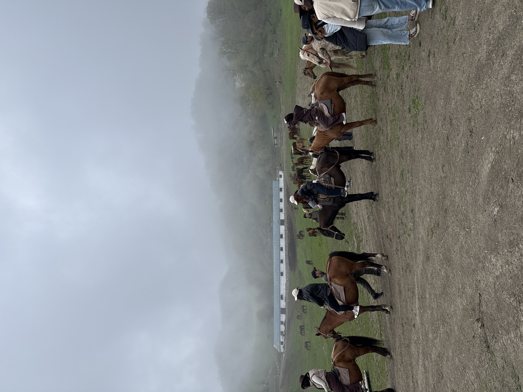 The image shows a group of people on horseback in a landscape near Haghartsin, Armenia. The setting is partly obscured by heavy fog and appears to be a grassy field adjacent to a series of white buildings. People are riding in a line. There are additional horses visible in the background. The terrain consists of dirt and grass with a hillside in the background.