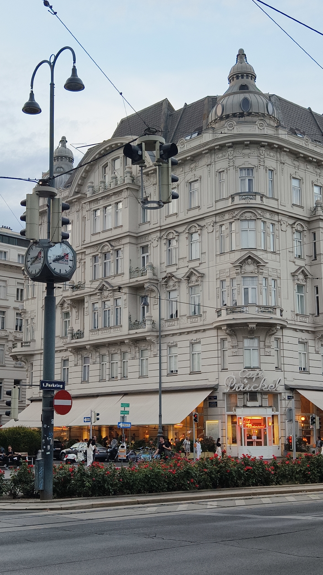 The image captures a street view in Vienna, Austria, featuring a prominent building on the right corner and a street lamp with a clock on the left. The architecture of the building is ornate, with classical detailing, a domed roof element, and multiple floors of windows. A sign on the building reads "Cafe Prückel." In front of the building, a row of red flowers lines the street, and several people are visible walking and sitting. The street lamp on the left is a functional element, holding traffic lights and a round clock face displaying "WIENER STÄDTISCHE." A "1. Parkring" street sign is also visible. The overall scene depicts an urban environment with pedestrian activity.
