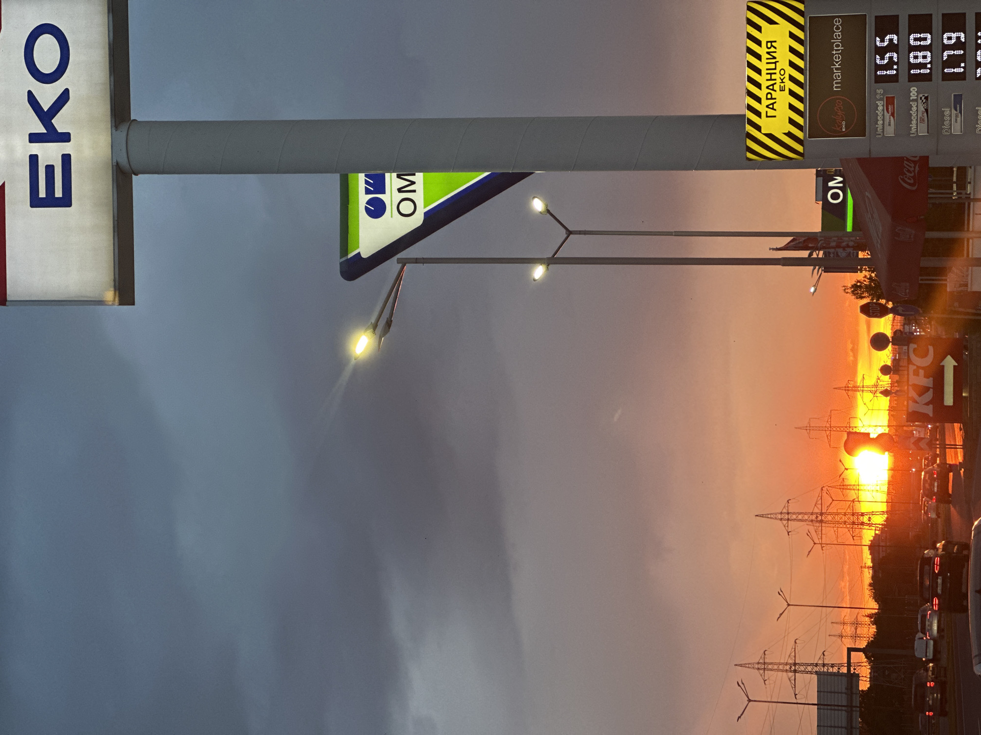 The image depicts a sunset view at a gas station in Sofia, Bulgaria. Prominent signs indicate the gas station is part of the EKO chain, and a smaller sign mentions "Kalypso marketplace." A KFC restaurant is also visible in the background. The sunset creates a vivid orange and red sky, with silhouettes of power lines in the foreground. Traffic, including cars, is visible on the road. The image also includes signage with fuel prices. The sky is a mixture of dark clouds and sunset colors.