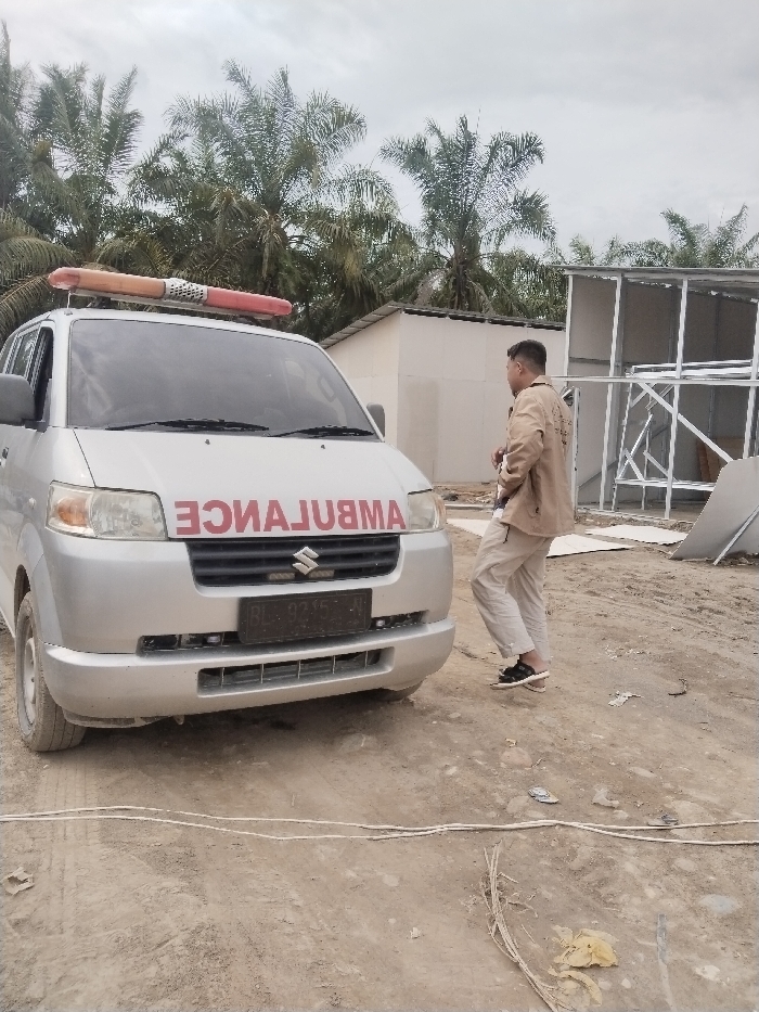 The image captures an outdoor scene featuring a silver ambulance van and a man, set against a backdrop of palm trees and construction elements.
In the foreground and center-left, a silver Suzuki van is clearly visible, functioning as an ambulance. The word "AMBULANCE" is prominently displayed in red, mirrored text across its front grille, indicating its emergency service role. A red and clear emergency light bar is mounted on its roof. The vehicle has a black Indonesian license plate, partially obscured but showing the characters "BL 9355" followed by what appears to be two more numbers. The "Suzuki" logo is also visible on the grille. The ambulance is parked on a dirt or sandy ground.
To the right of the ambulance, a man stands facing away from the camera, looking towards the right side of the frame. He is wearing a light brown or tan jacket and light-colored, possibly beige or white, pants. He has short, dark hair and is wearing dark-colored sandals. His right arm is slightly bent, and his left hand is not fully visible.
The background features several tall palm trees with green fronds, suggesting a tropical or subtropical environment, possibly an agricultural area or a coastal region. Behind the ambulance and the man, there are a few structures. A white, box-like building is seen behind the ambulance. Further to the right and slightly behind the man, there is an unfinished or partially constructed building made of light-colored metal framing, possibly a pre-fabricated structure, with some white paneling nearby.
The ground is unpaved, consisting of light brown dirt or sand, with tire tracks visible near the ambulance's front wheels. Scattered debris, including some white cords, dry leaves, and what appears to be a crumpled plastic bag, are on the ground in the foreground. The sky is overcast and light gray, indicating a cloudy day. The overall impression is one of a functional scene in a developing or semi-rural area, possibly associated with a worksite or a remote community.