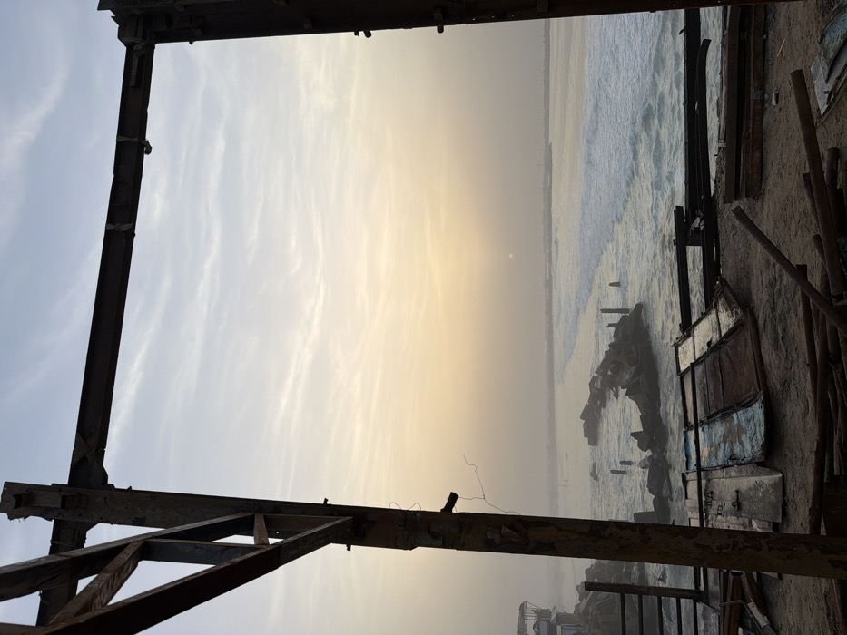 A hazy sky meets the ocean horizon framed by rusty, dilapidated metal structures. The sky shifts from light blue to yellow where the sun is positioned over the distant ocean. Waves crash against a rocky outcrop, and several weathered posts stand in the water. Debris and wooden poles litter the sandy foreground, creating a stark contrast between the industrial decay and the natural seascape.
