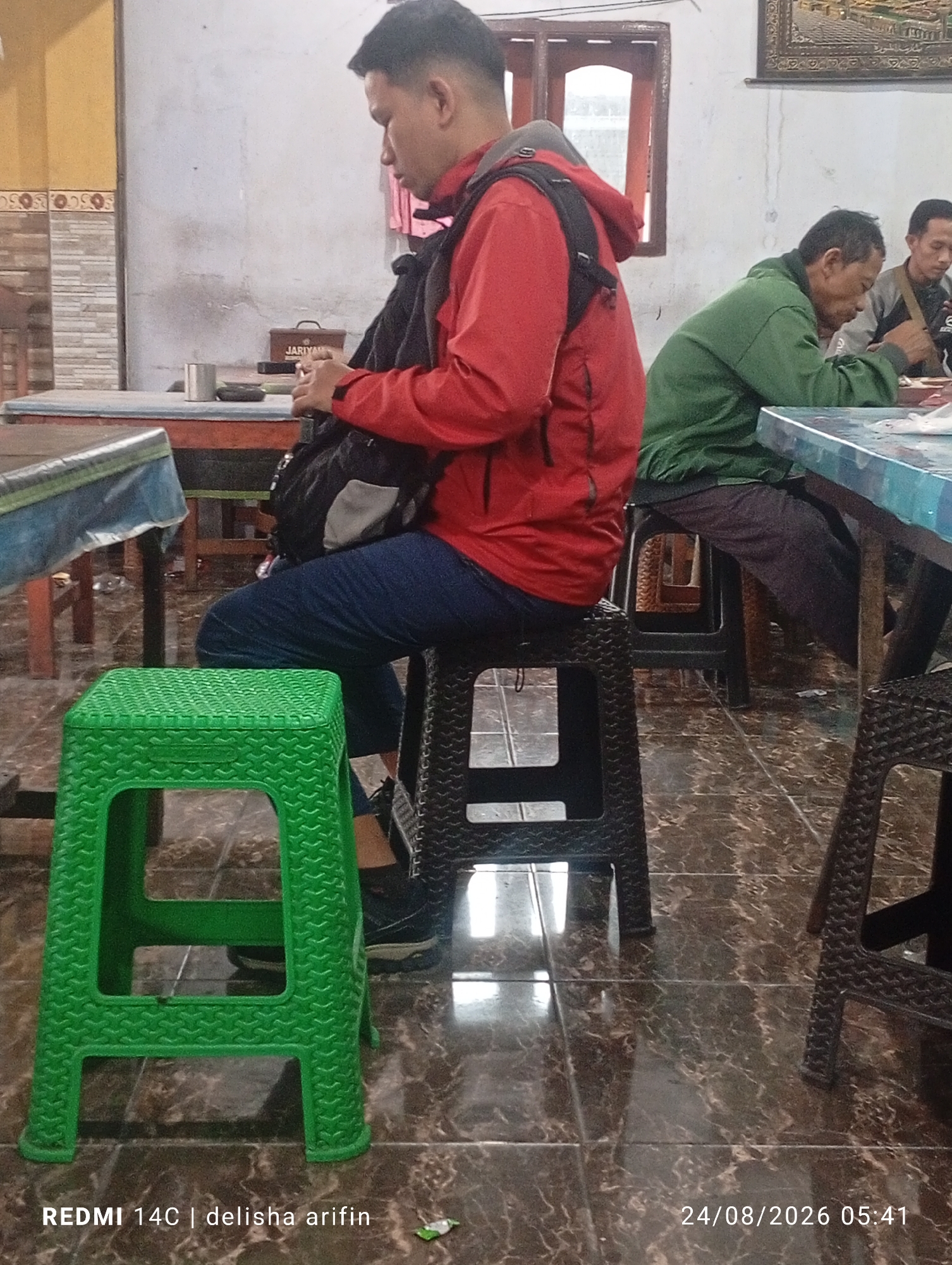 The image depicts a casual indoor scene, possibly a cafe or eatery, in Gambir Kuning, Indonesia. In the foreground, a young man wearing a red jacket and blue jeans is seated on a dark-colored plastic chair. He has a black backpack slung over his shoulder. To his left, a bright green plastic stool is positioned. In the background, two other men are seated at a table. One man, wearing a green jacket, is looking down at his hands. Another man to his right, partially visible, is also looking down. The environment appears to be a simple interior with tiled flooring and a light-colored wall. A table with a blue and white patterned surface is visible in the mid-ground, and another wooden table is behind the man in the red jacket. The overall lighting suggests it is daytime, though the light is somewhat dim. The date and time stamp at the bottom right reads "24/08/2026 05:41", and the bottom left reads "REDMI 14C | delisha arifin".