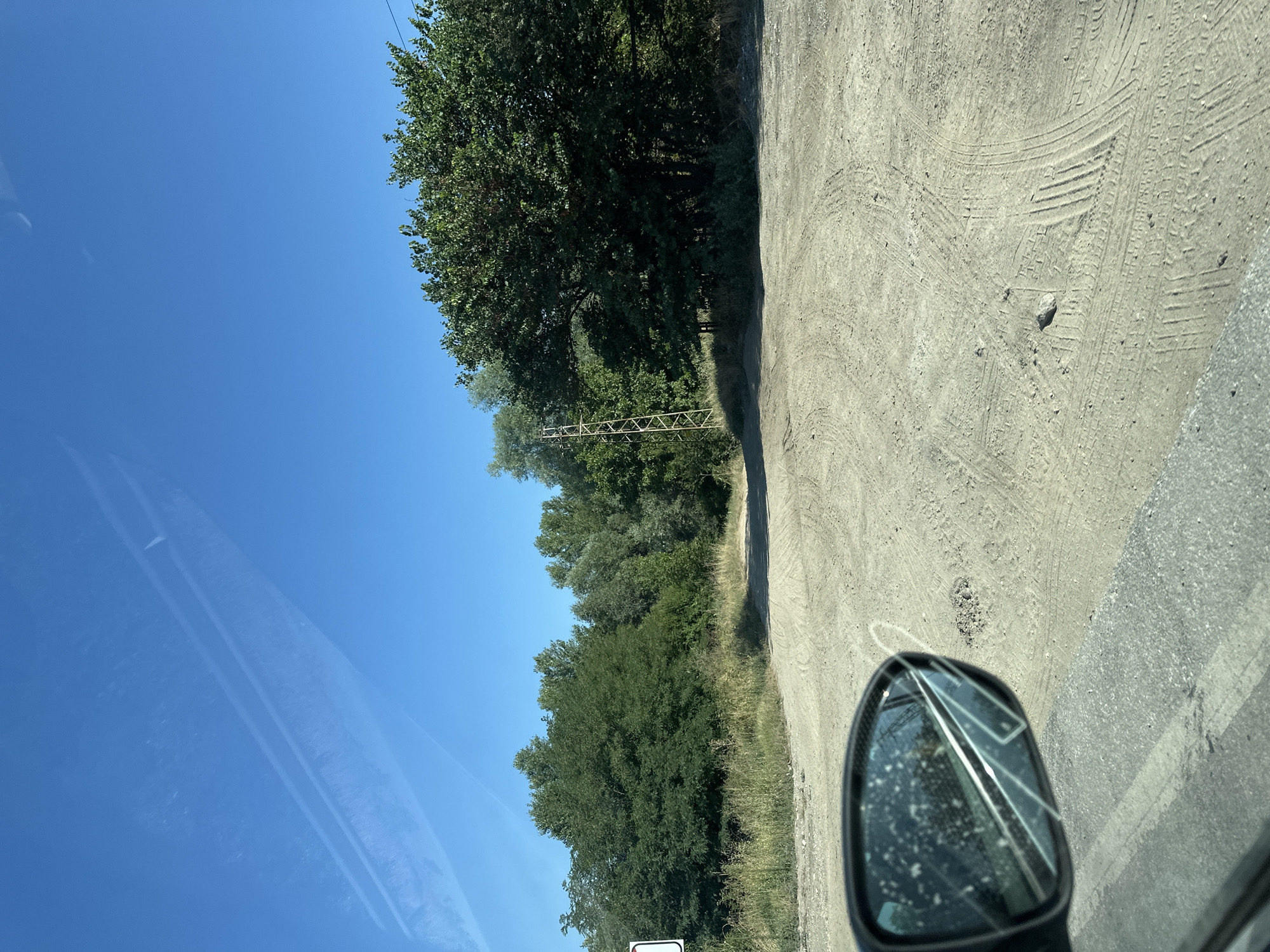 The image depicts a dirt road leading into a wooded area under a clear blue sky. A side mirror of a vehicle is visible in the lower left corner, reflecting the surroundings. In the distance, a metal utility pole stands amongst the trees. Tire tracks are imprinted on the dusty road, indicating recent vehicle activity. The scene appears to be located in the vicinity of Yoakim Gruevo, Bulgaria.