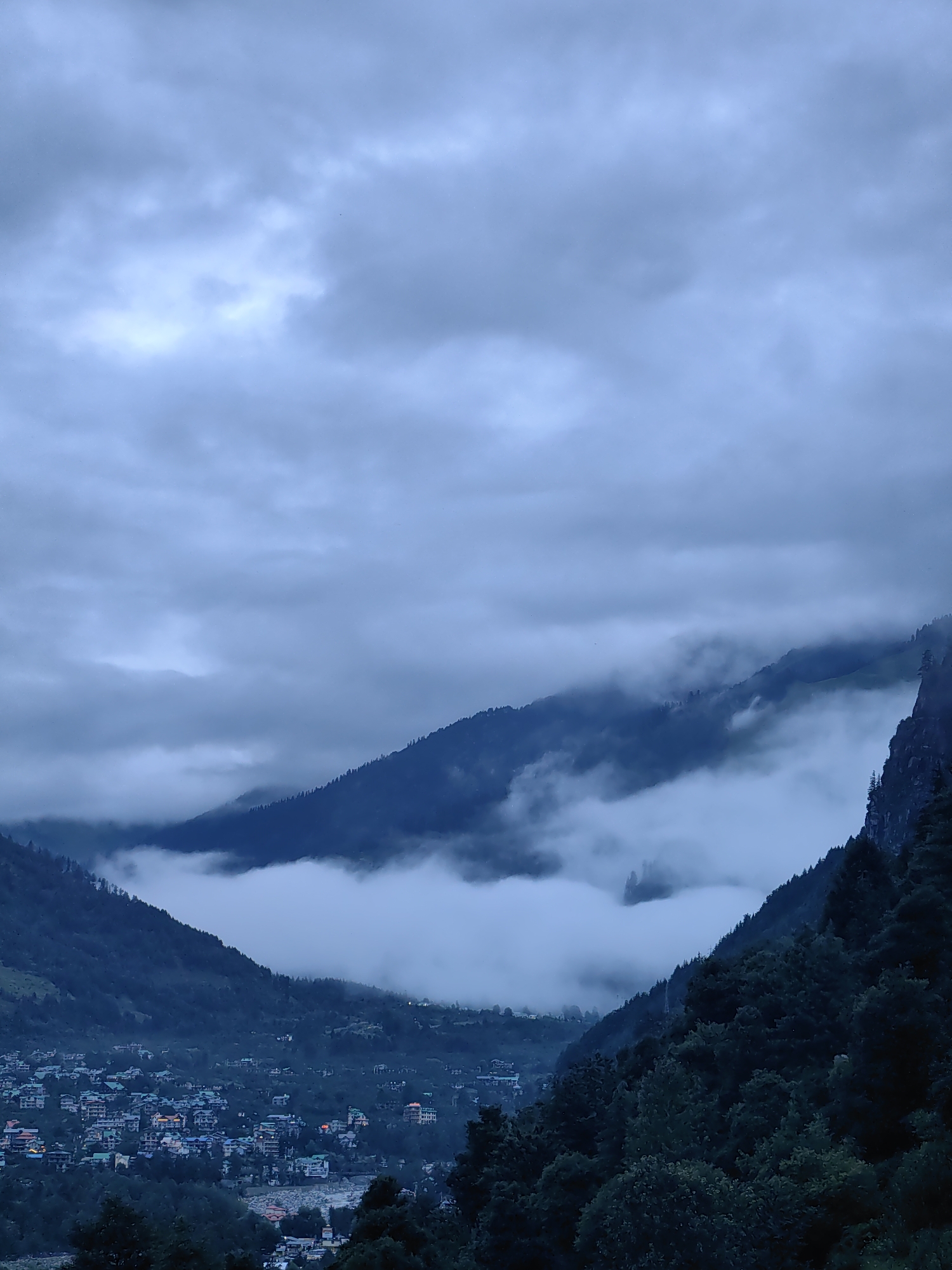 The image captures a dramatic, overcast day in the mountainous region of Vashisht, India. The scene is dominated by a valley filled with dense fog and low-hanging clouds, which partially obscure the slopes of the surrounding mountains. The mountains themselves are covered in dark green foliage, with the tips of some trees visible above the mist.
Nestled within the valley, a small town is visible, characterized by a cluster of buildings with multi-colored roofs. These buildings appear to be of varying sizes and styles, indicative of a residential area. Some lights are visible within the buildings, suggesting it is either early morning or evening.
The sky above is a uniform gray, with thick clouds that create a moody and atmospheric effect. A brighter patch of light in the upper left quadrant of the sky suggests a potential break in the clouds or sunlight struggling to penetrate. The overall impression is one of a serene, yet slightly somber, mountain landscape. There are no people or specific actions depicted in the scene.