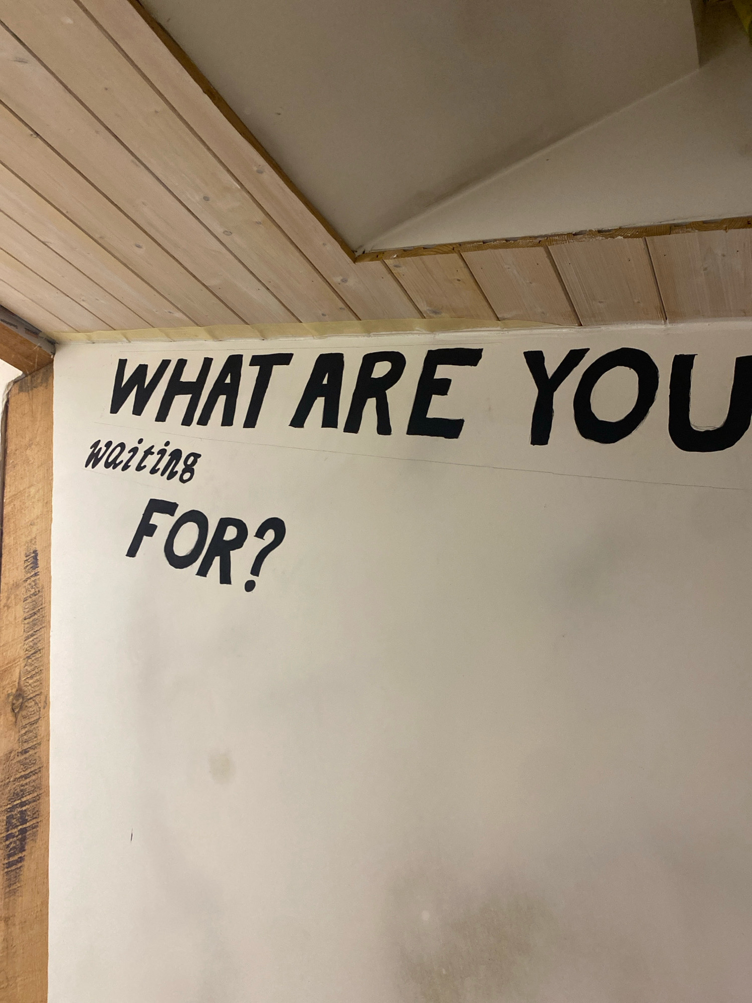 The image shows a white wall with black lettering. The top line of text reads "WHAT ARE YOU". Below this, to the left, is the word "waiting" written in a smaller, script font. Below "waiting" and centered on the wall is the word "FOR?" in a large, bold font, followed by a question mark. The wall is located in Mazlauki, Latvia. A wooden beam is visible on the left side of the image, and wooden planks form a ceiling overhead, angled downwards. The wall appears to be part of an interior space, possibly a workshop or garage.
