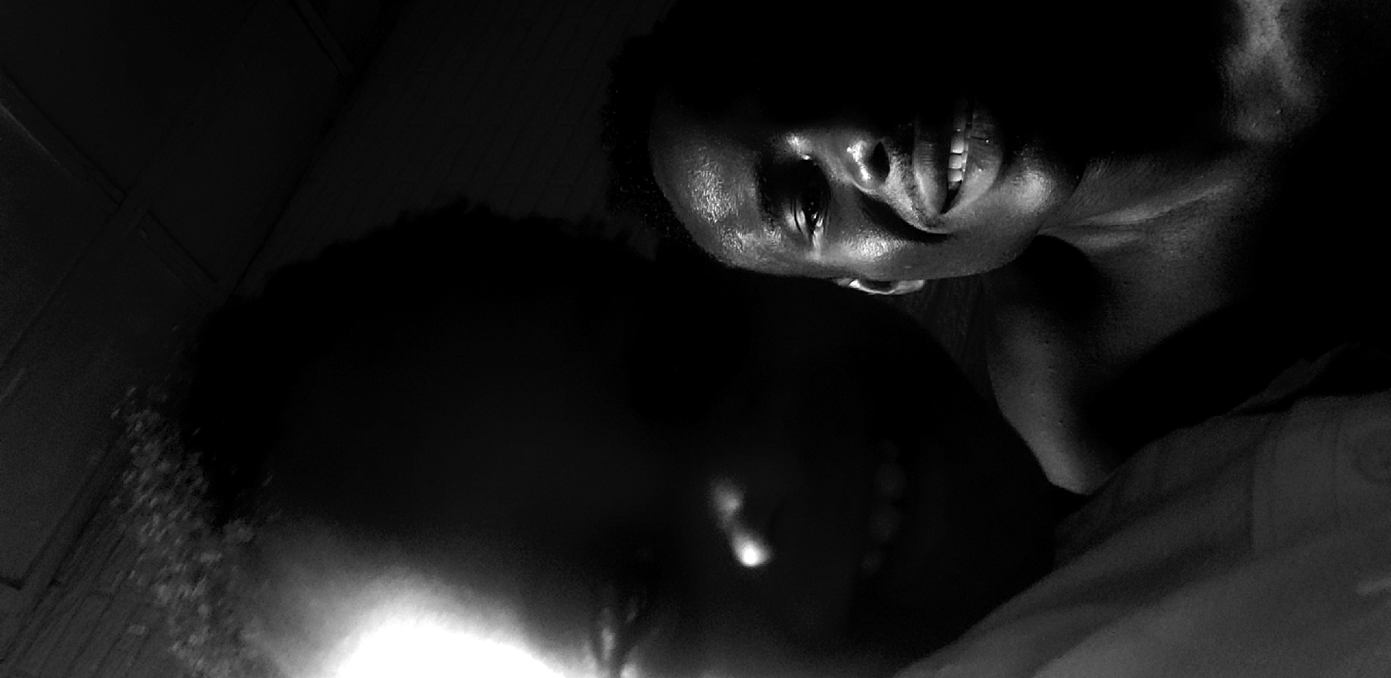 This is a black and white, low-light, close-up image of two individuals, likely a couple, in an intimate setting. One person's face is partially visible, turned upwards, with their mouth open in a smile or laugh. Their skin appears to glisten, possibly from sweat or moisture. The lighting is dramatic, casting deep shadows and highlighting facial features. The other individual's face is mostly obscured by darkness, with only a hint of their features visible. The overall mood is suggestive of a private and possibly romantic moment. The background is dark and indistinct, offering no clear clues about the location, other than the provided context of Narayi, Nigeria. There is no visible text in the image. The time of day is not discernible due to the low light, but the intimacy of the scene might suggest nighttime.