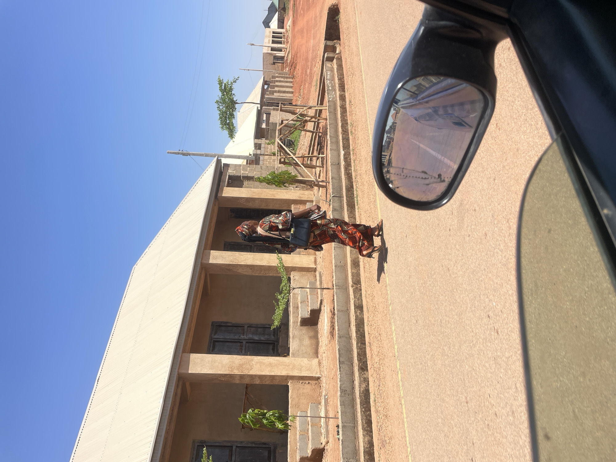 The image depicts a street scene in Jos, Nigeria, viewed from inside a vehicle. A woman in traditional African attire, including a patterned dress and headwrap, walks along the roadside, carrying a dark-colored bag. A two-story building is positioned to her left, with small trees planted along its facade. Further down the road are other buildings, some under construction. The sky is clear and blue. Part of the vehicle's side mirror is visible in the foreground, reflecting the scene behind the vehicle. The road is paved and appears mostly empty.