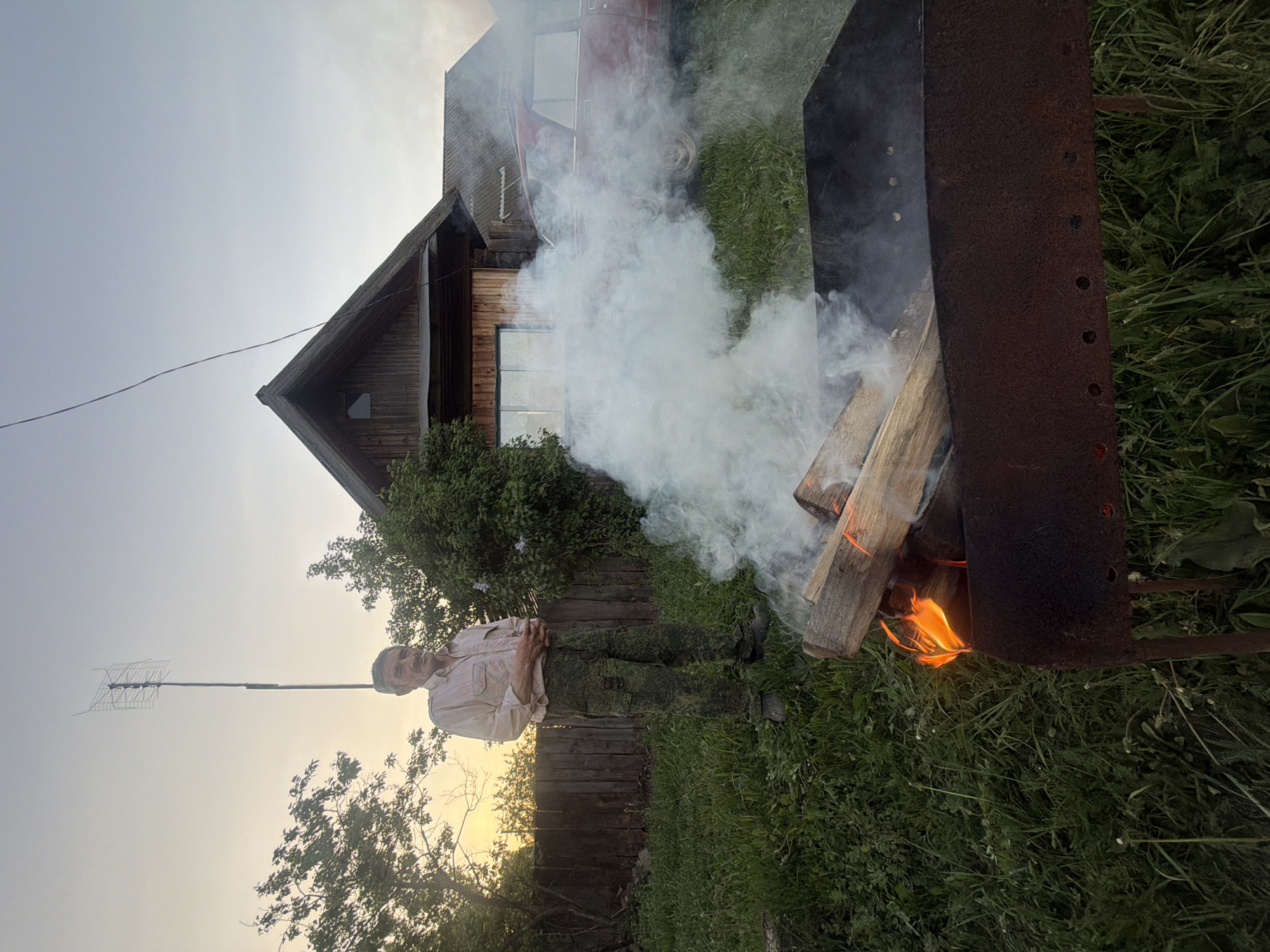 A photograph taken in what appears to be Dubrovo, Russia, shows a man standing near an active fire pit. The fire pit contains burning logs emitting flames and thick smoke. The man, dressed in light-colored shirt and camouflage pants, is standing near a wooden fence. In the background is a house and a parked car. An antenna is visible in the sky. The ground is covered in green grass and vegetation. The photograph has a warm, evening light.