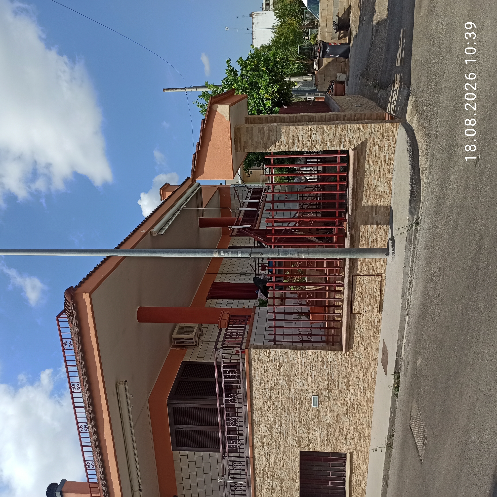 The image depicts a residential building with a distinct terracotta-colored roof and orange exterior walls. The building features a balcony with a decorative metal railing and a stone-tiled lower facade. In front of the building, a red metal fence encloses a small area with potted plants. A street runs in the foreground, with a sidewalk and a storm drain visible. The sky is blue with scattered white clouds. A tall metal pole stands on the sidewalk. In the background, trees and other buildings can be seen. The date and time stamp "18.08.2026 10:39" is visible in the lower right corner. The scene appears to be a quiet residential street in Bitonto, Italy, likely during daytime.