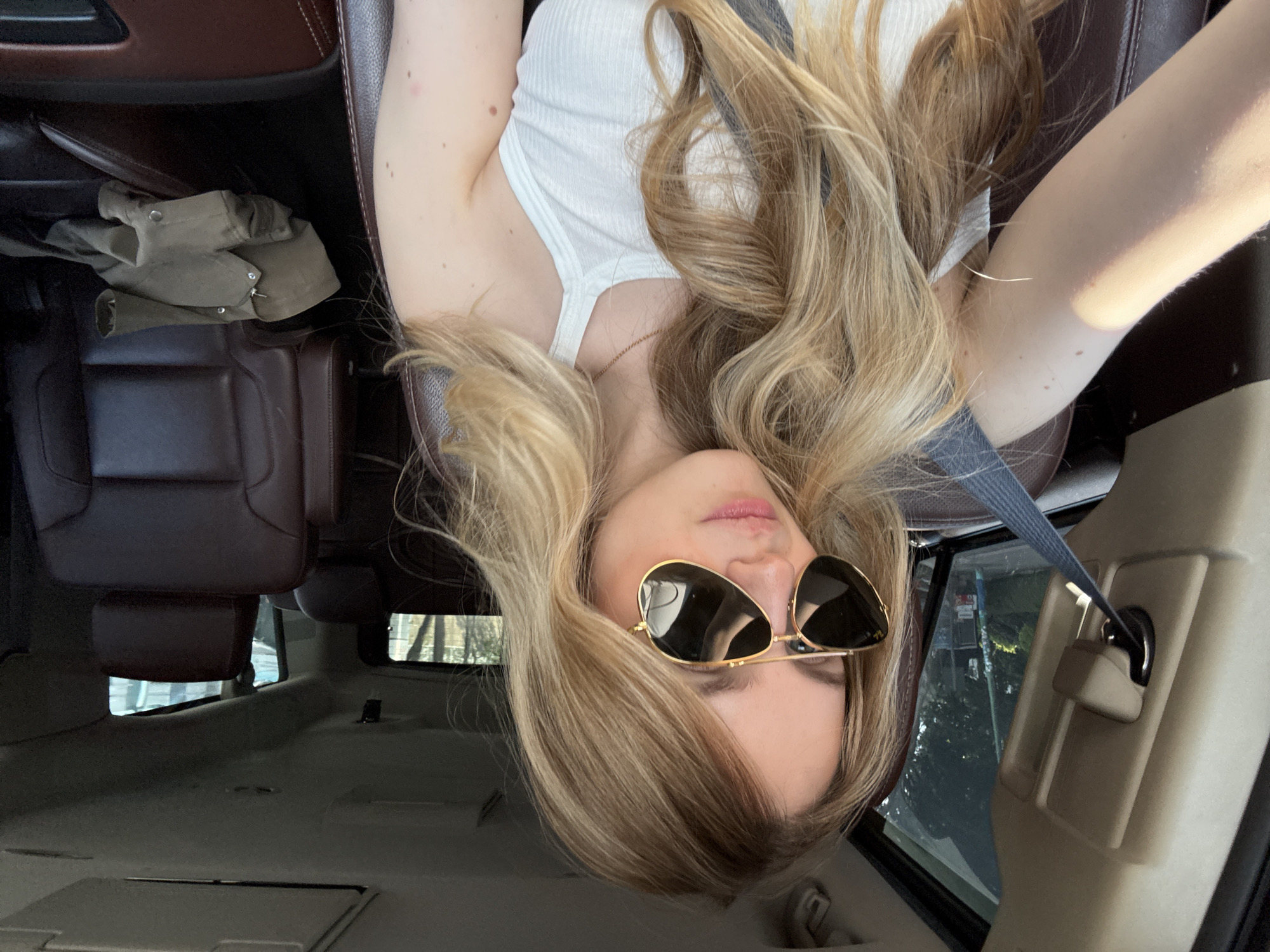 A blonde woman wearing sunglasses and a white tank top is lying in a dark-brown leather car seat, fastened with a grey seat belt. A tan jacket sits on the adjacent seat, and the vehicle's interior is visible, including seats and the back area. Outside the windows, trees are visible, suggesting an urban or suburban environment. The image does not provide information to ascertain that this is Mexico City, Mexico.