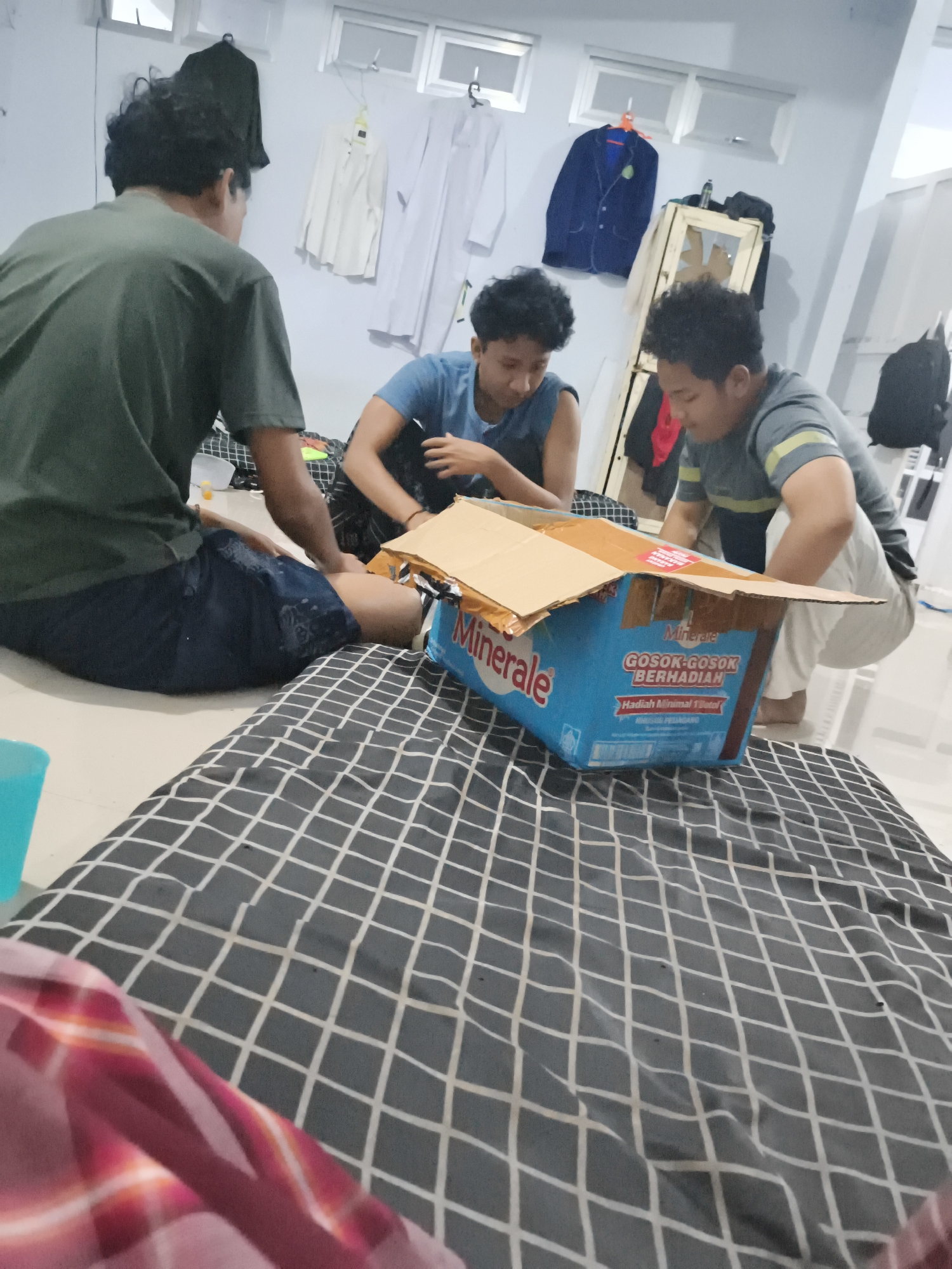 In Ketawang, Indonesia, three individuals are gathered around a large blue cardboard box on a patterned mat. One person, in the foreground and facing away from the camera, wears a dark green t-shirt and dark shorts. To their left, another person in a light blue t-shirt is seated, looking down at the box. The third person, wearing a grey and yellow striped t-shirt and white pants, is crouched next to the box, interacting with it. The box is open and appears to contain items. The environment is indoors, with white walls, windows, and clothing items hanging on a rack in the background.