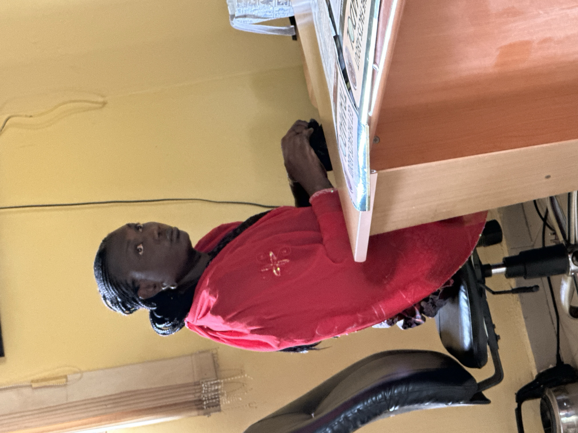 A medium shot captures a Black woman with braided hair, wearing a red dress and a silver earring, seated at a wooden desk in an office. She is looking to her right with a contemplative expression. Her hands are resting on the desk, appearing to be engaged with something out of frame. The desk surface has several documents or papers laid out. To the left of the woman, a black office chair with a leather seat is visible. The background features a yellow-painted wall with a window on the left, partially covered by sheer curtains. The scene suggests an indoor office environment in Kaduna, Nigeria.